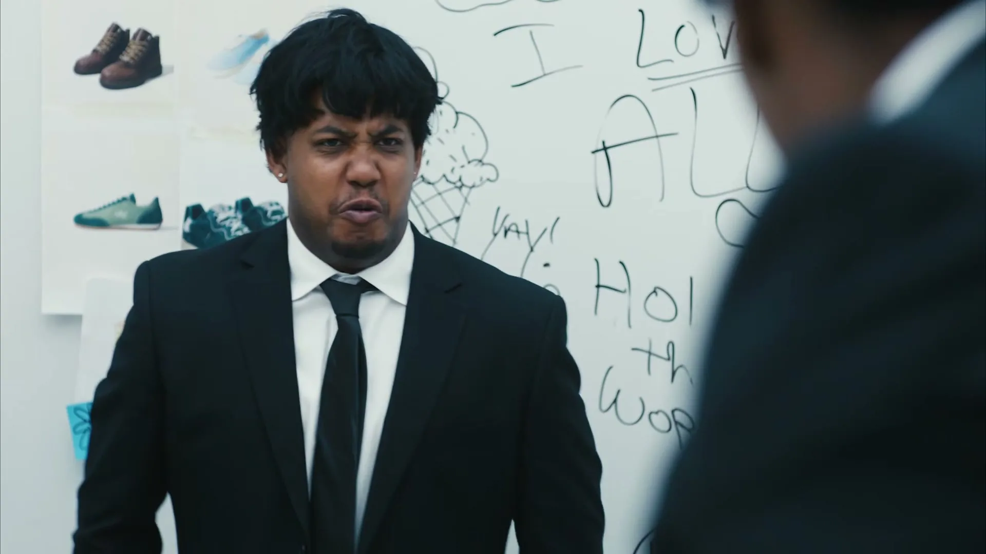 A young man in a black suit and tie looks directly at the camera with a serious expression, standing in front of a whiteboard with drawings and text.