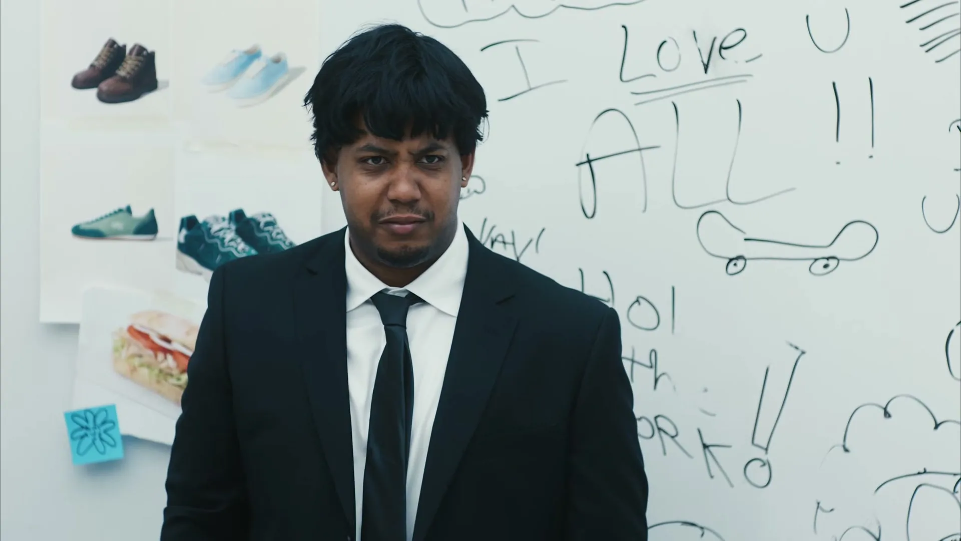 A man with dark hair and a black suit stands in front of a whiteboard covered in doodles and text, looking directly at the camera with a slight frown.