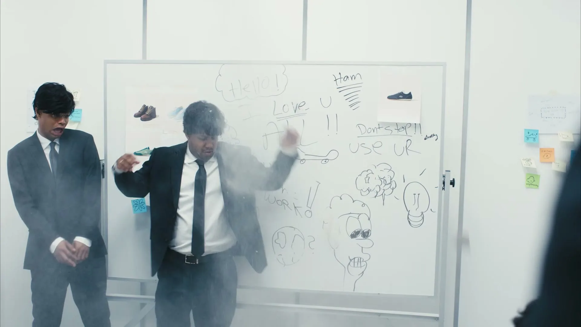 Two men in black suits stand in front of a whiteboard, one gets hit by a cloud of white powder, in an office.