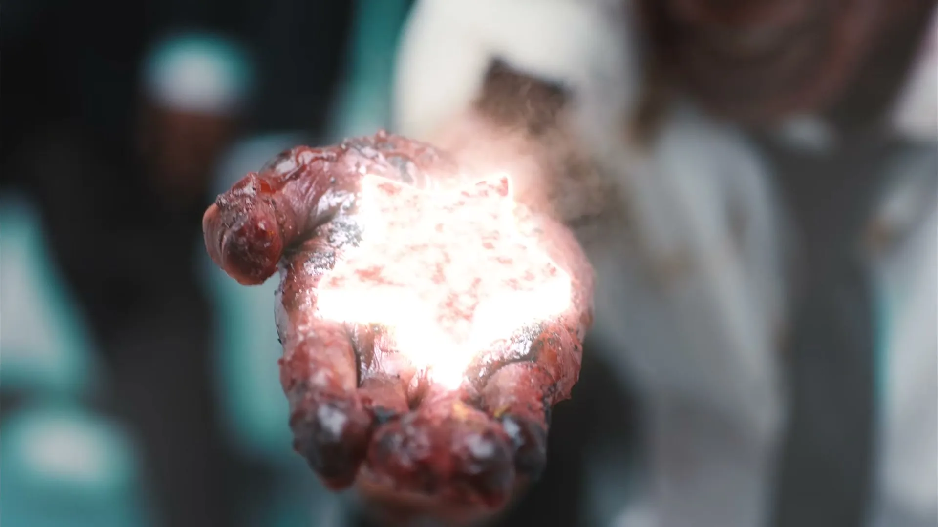 A person's hand, covered in what appears to be blood and grime, holds a glowing, star-shaped object that emits light and smoke.