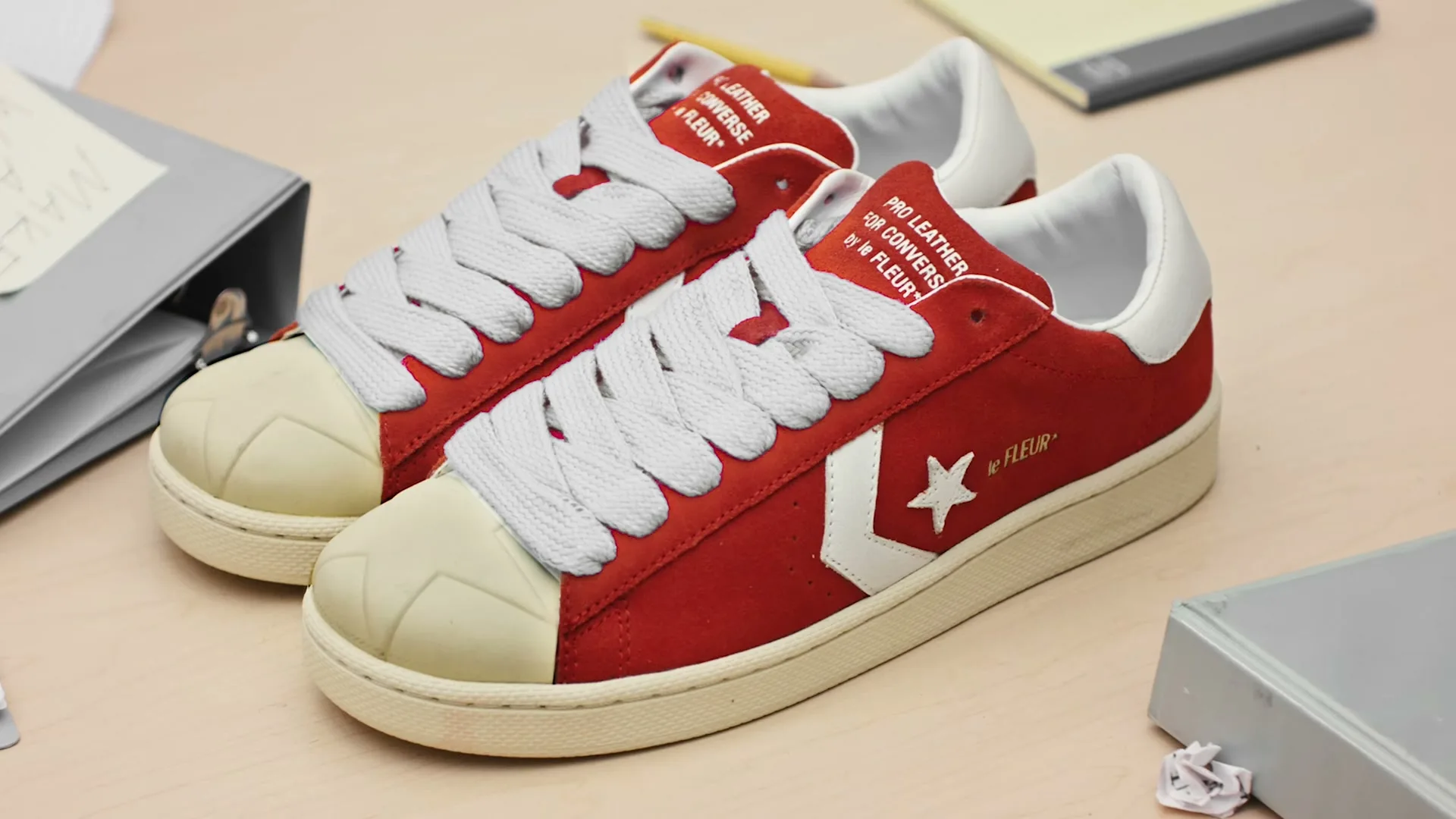 A pair of red Converse Pro Leather by le FLEUR* sneakers with white laces and toe caps are displayed on a light wooden desk with office supplies.