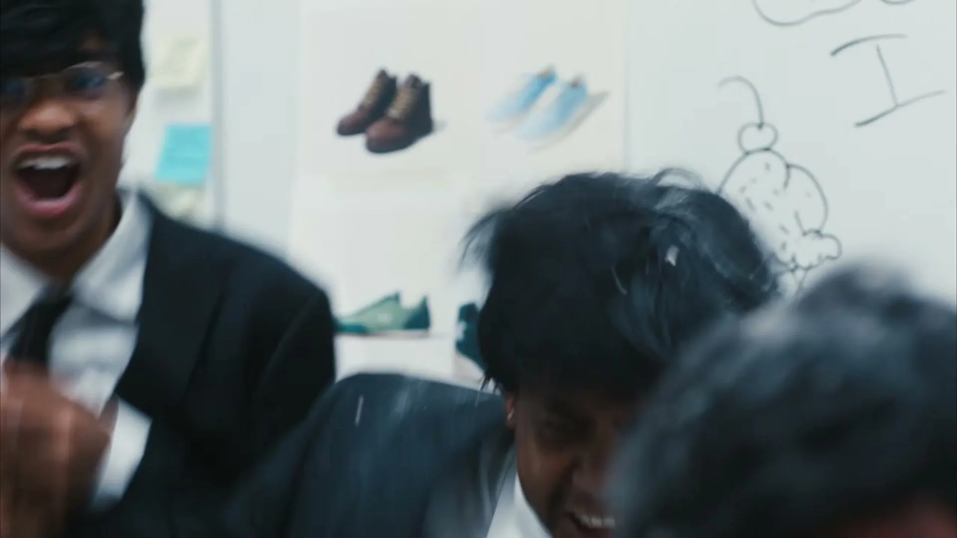 Two young men in black suits and ties are covered in white powder, one with glasses is yelling, the other is grimacing, in a white room with drawings on the wall.