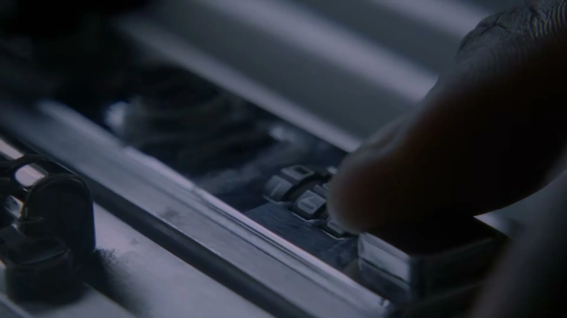 A person's finger adjusts the combination lock on a silver suitcase, revealing the numbers, in a close-up shot.