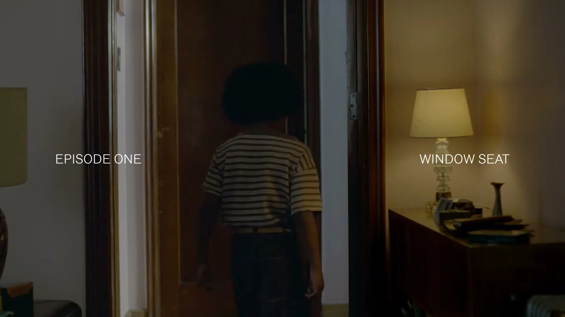 A young person with an afro, wearing a striped shirt and dark pants, stands in a doorway, looking into a dimly lit room with a lamp.