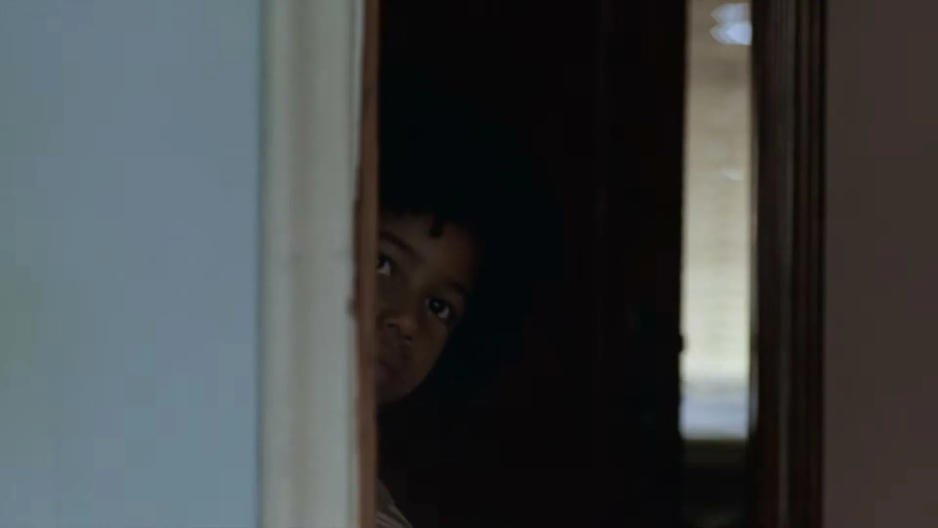 A young child with dark curly hair peeks out from behind a door frame, looking shyly into the room.