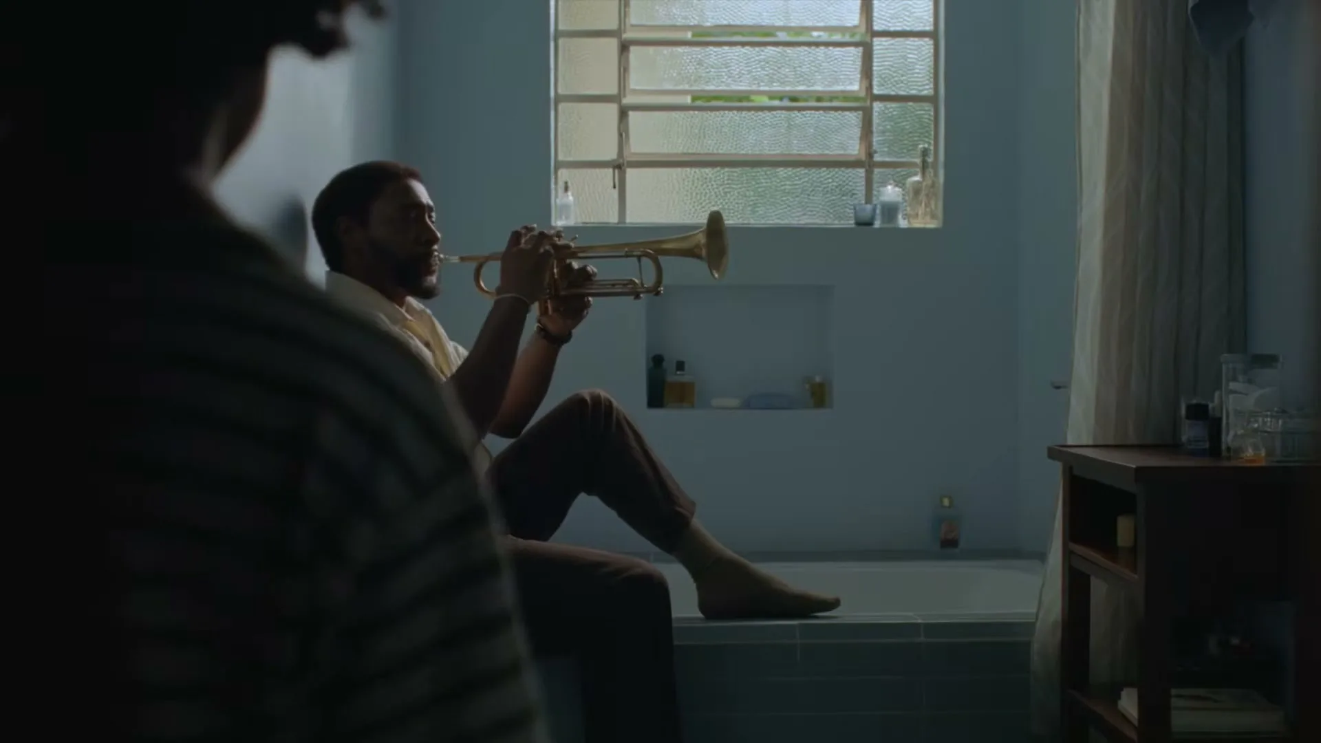 A man in his 30s sits on the edge of a bathtub, playing a trumpet, while another person watches from the foreground in a bathroom.