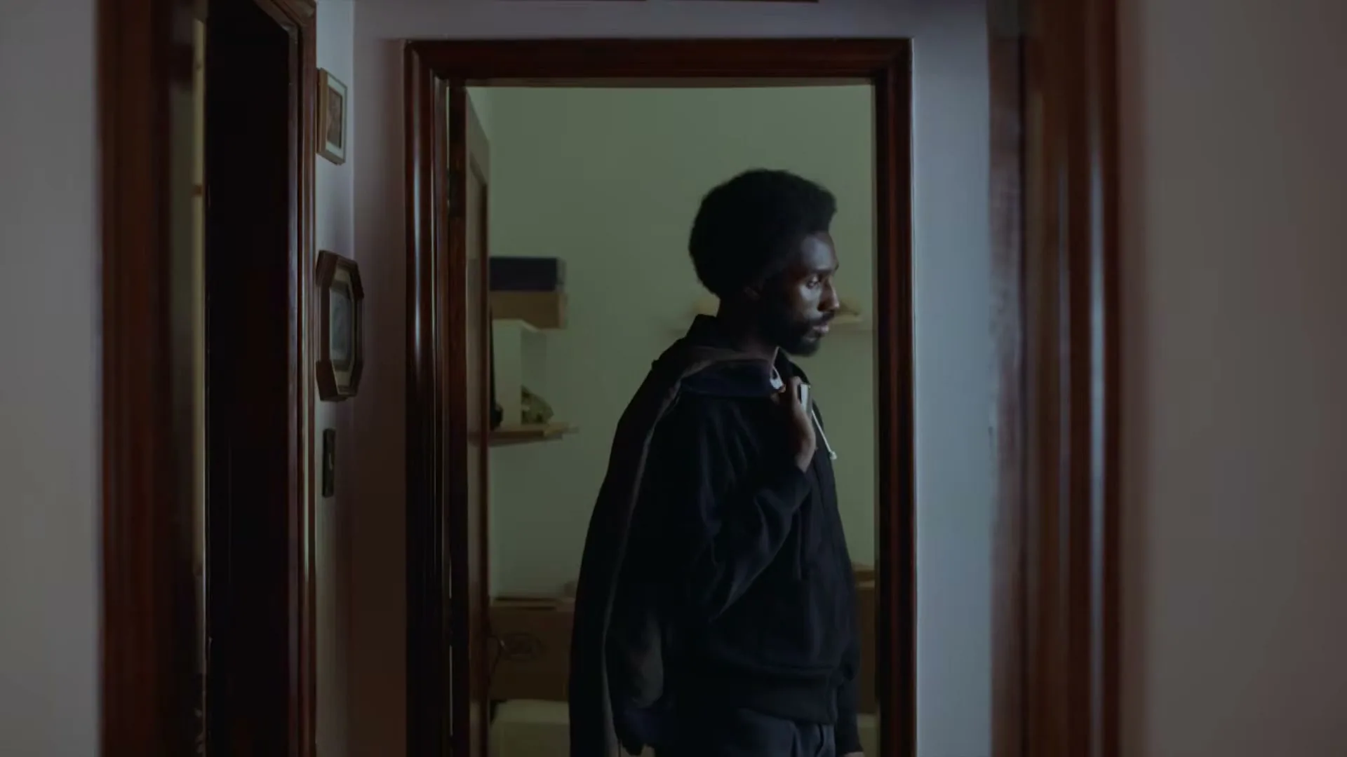 A man with an afro and beard, wearing a dark hoodie and carrying a jacket, stands in a doorway looking to the right.