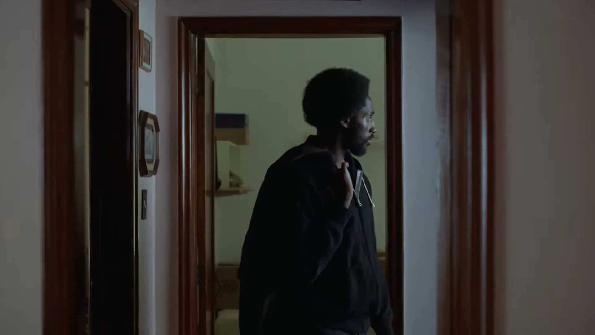 A young Black man with an afro, wearing a black hoodie, stands in a doorway, looking over his shoulder in a dimly lit hallway.