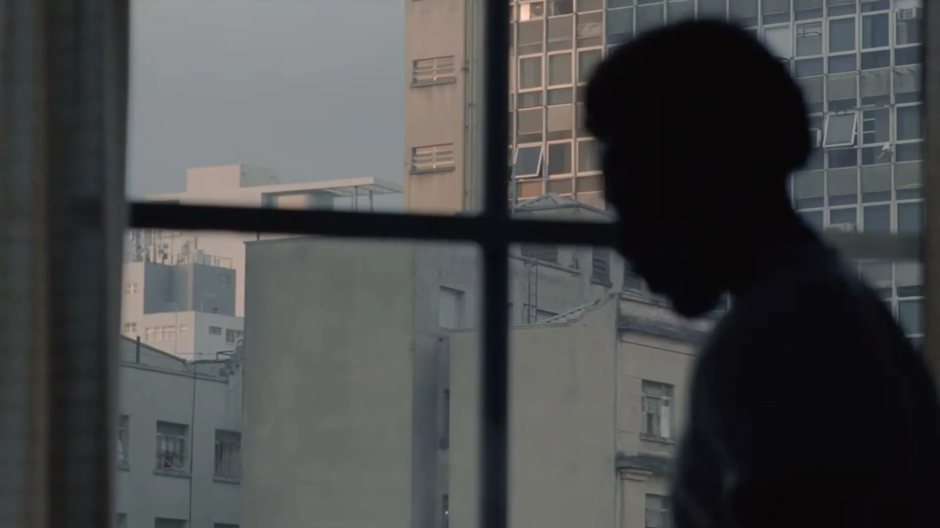 A silhouetted man stands by a window, looking out at a city skyline with buildings reflecting the golden hour light.