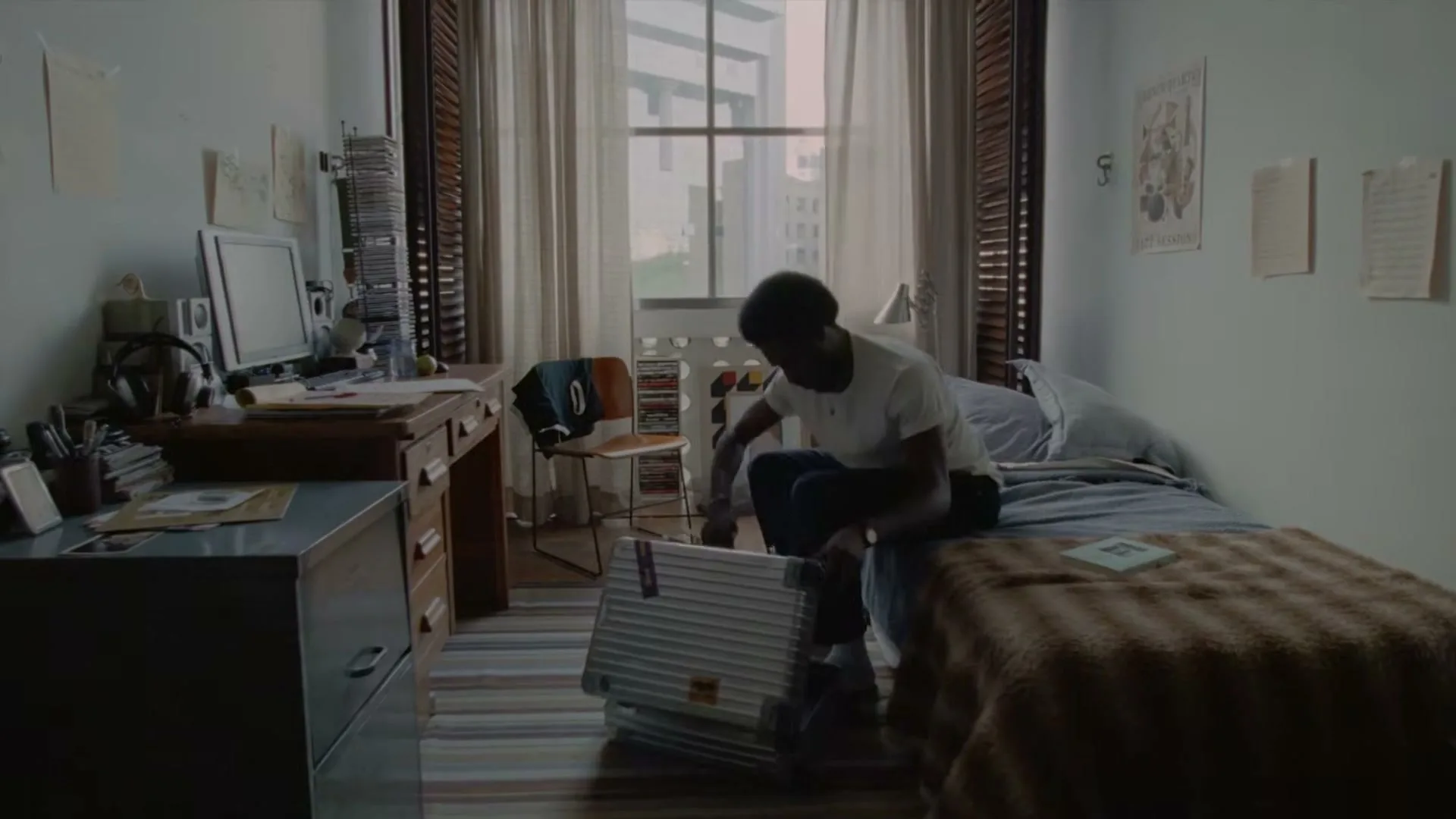 A young Black man with an afro sits on his bed, packing a silver Rimowa suitcase in a sunlit bedroom.
