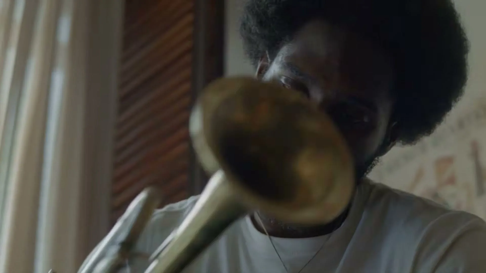A young Black man with an afro and beard, wearing a white t-shirt, looks down at a brass instrument he is holding in an interior room.