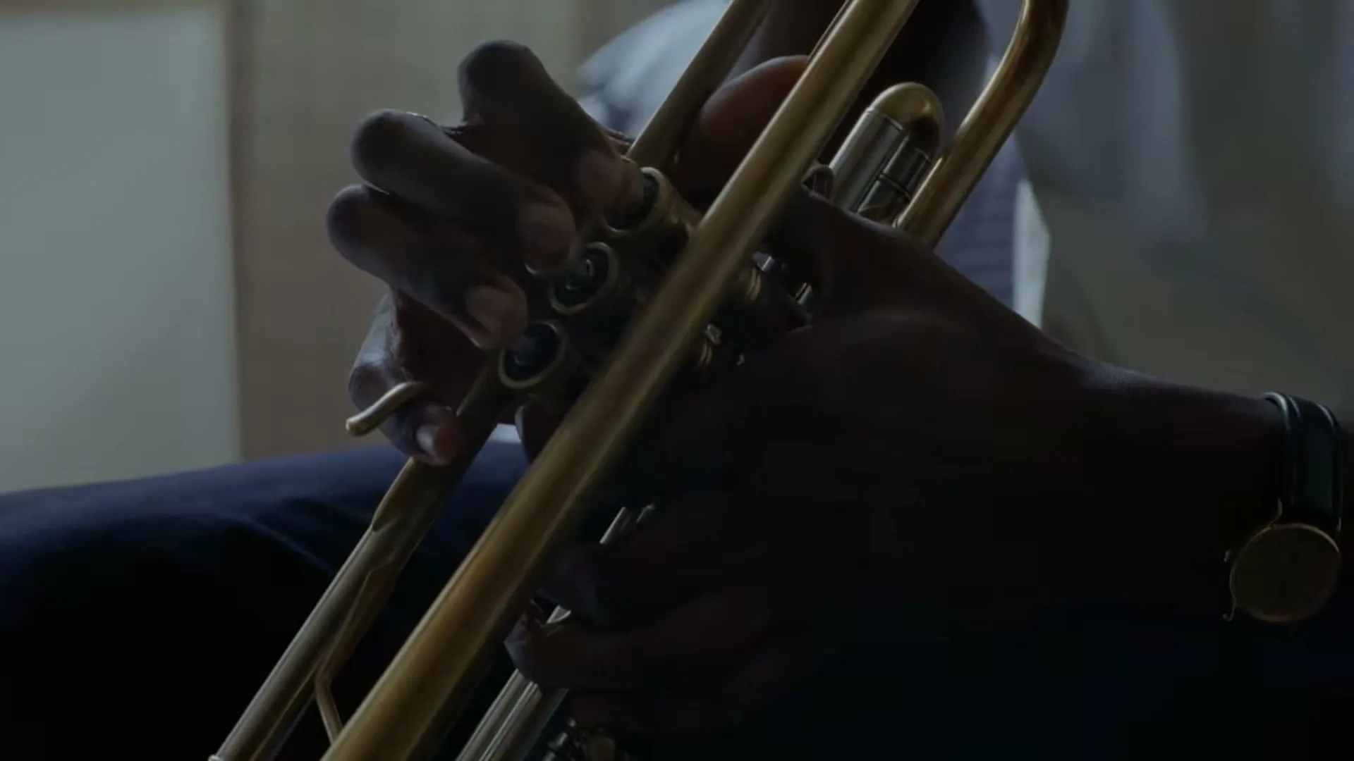 A person's dark-skinned hands are shown in a medium closeup, pressing the valves of a golden trumpet, in an interior setting.