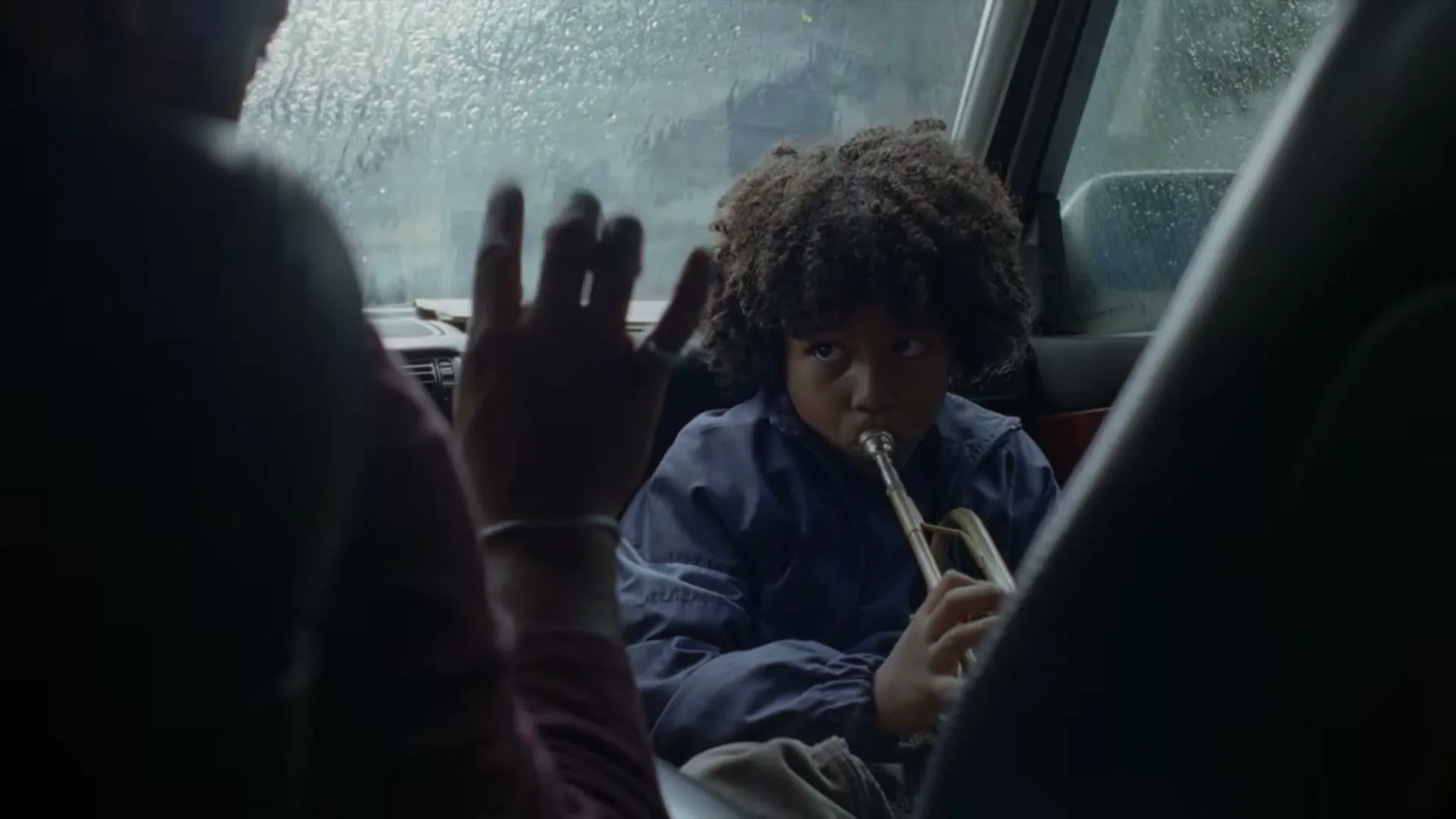 A young boy with curly hair plays a trumpet in the back seat of a car, while an adult's hand makes a peace sign in the foreground.