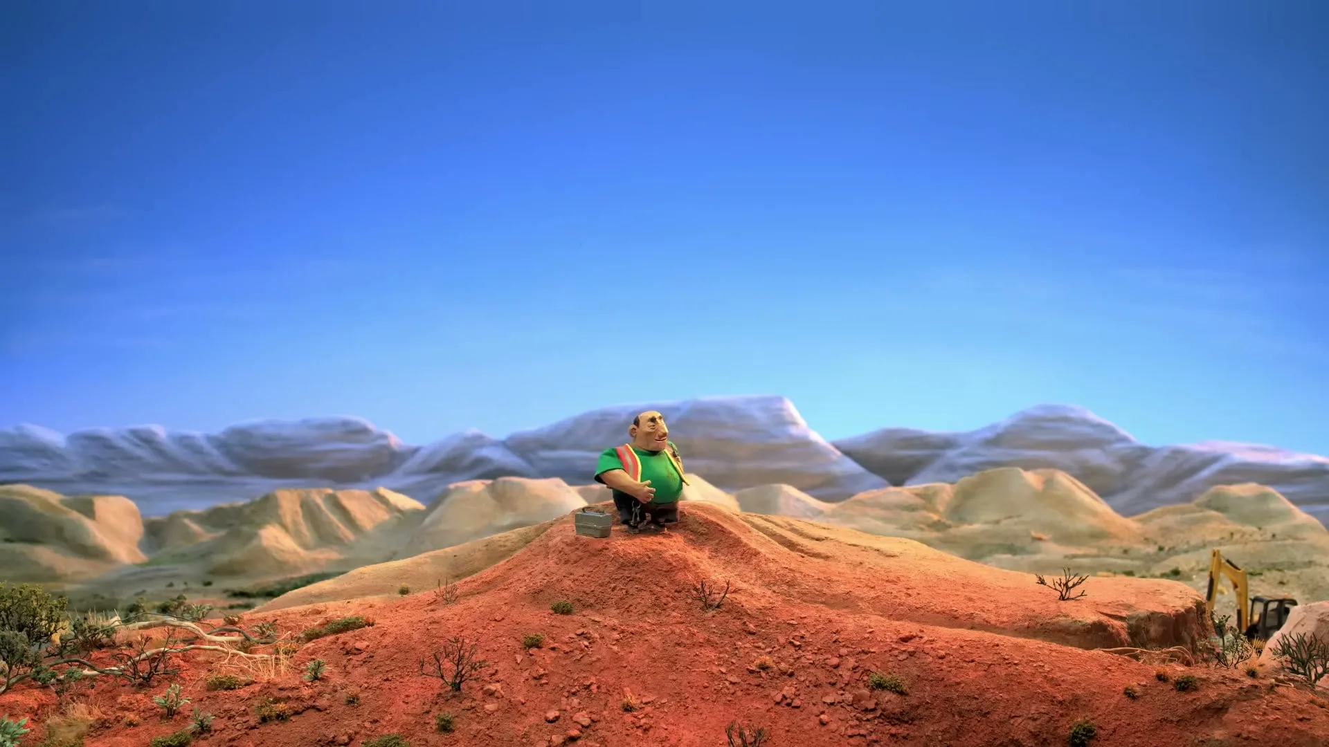 A stop-motion animated man in a green shirt and overalls sits on a small hill in a vast desert landscape, looking around.