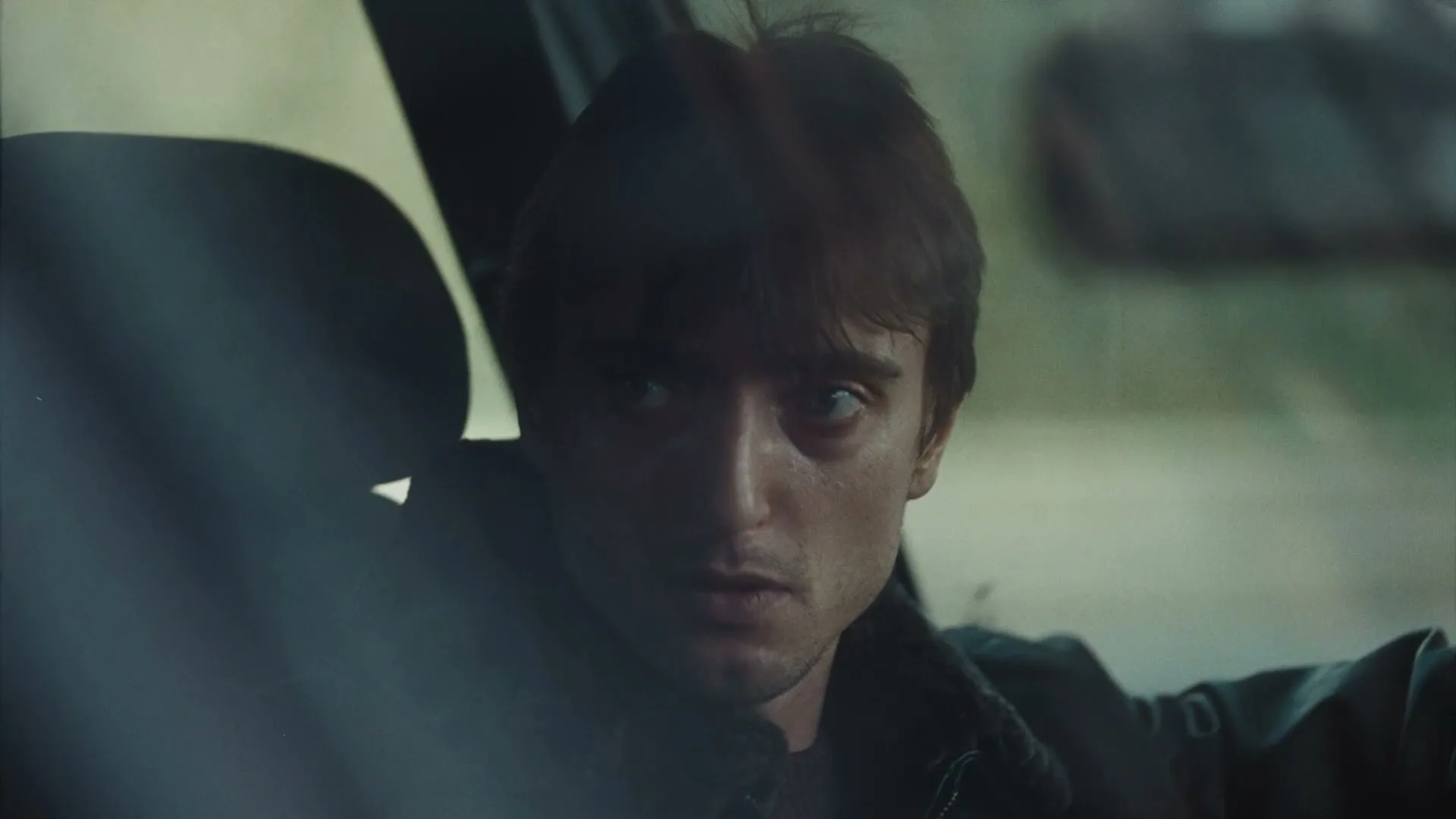 A young man with dark hair and a dark jacket sits in a car, looking around nervously, with a blurred background visible through the window.