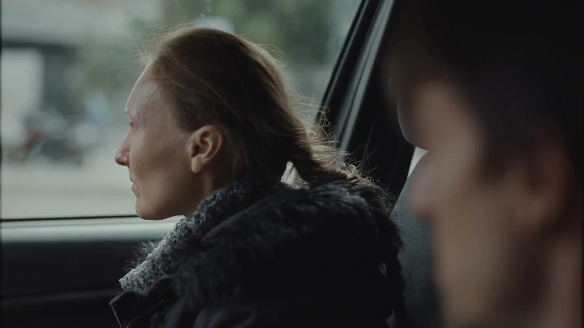 A woman with braided hair sits in the passenger seat of a car, looking out the window with a serious expression, as a blurred figure sits in the driver's seat.