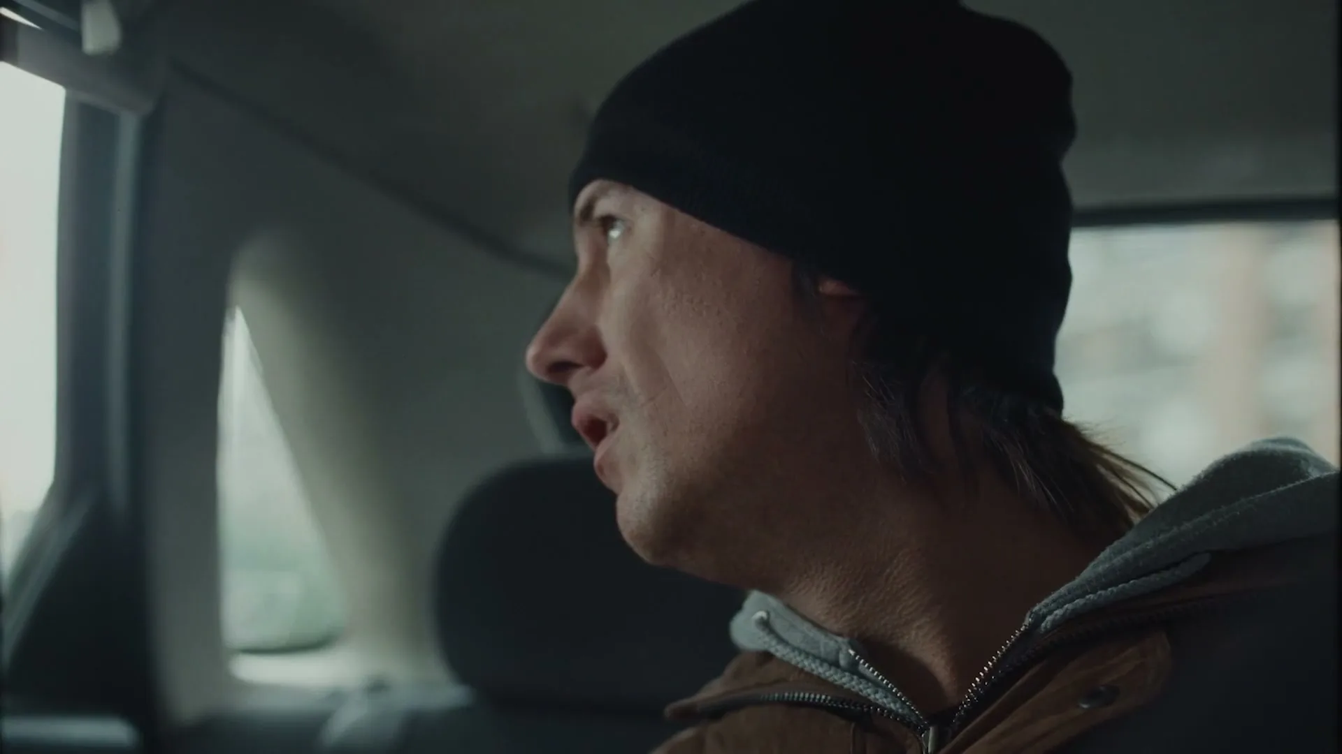 A young man with a black beanie and brown jacket looks out the car window, then turns to speak to someone off-screen in a car.