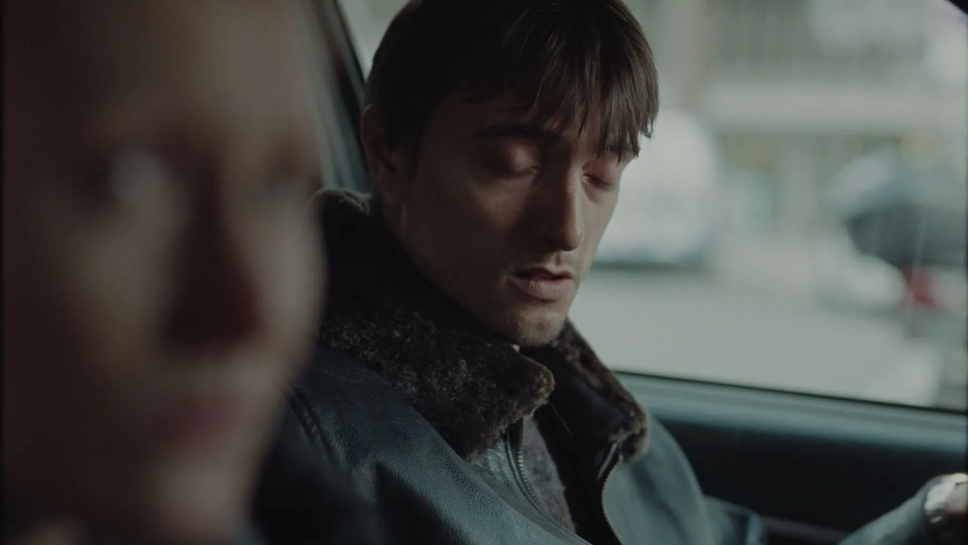 A man with dark hair and a leather jacket sits in a car, looking down then turning to speak to someone out of frame, with a blurred person in the foreground.