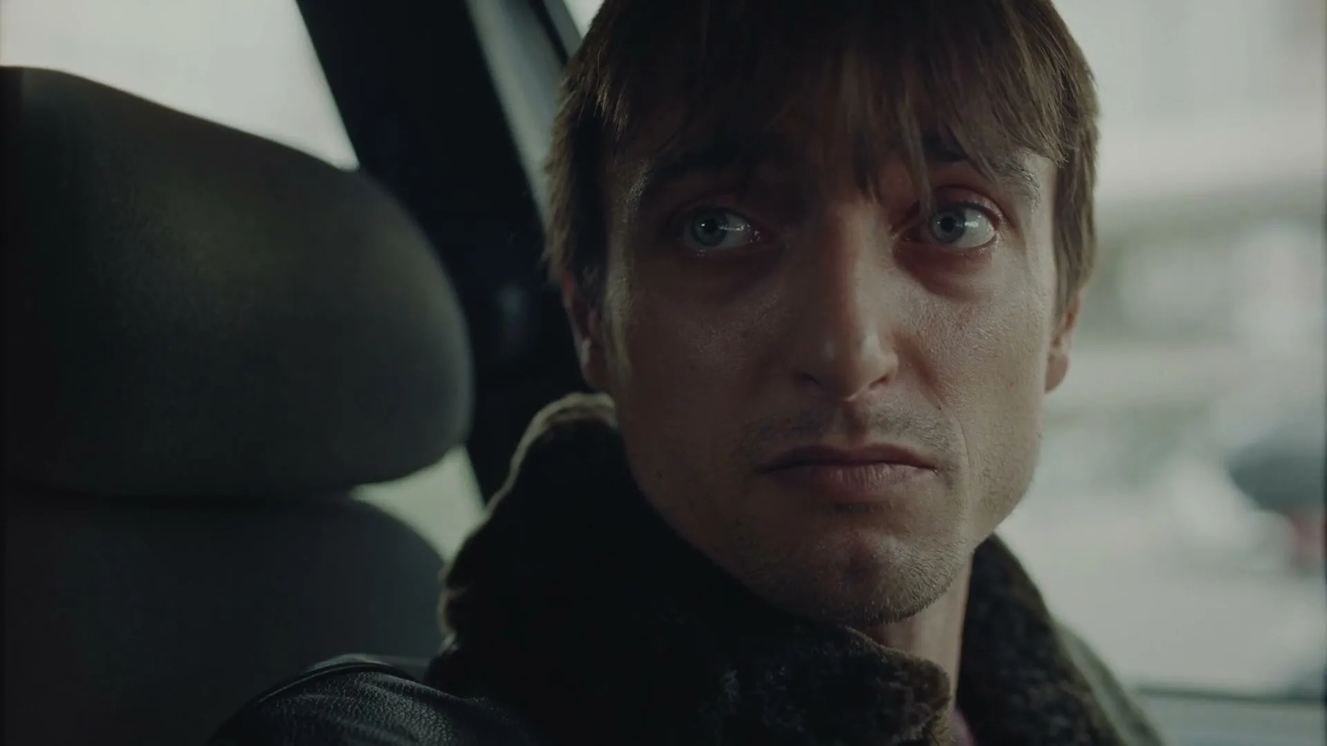 A man with shaggy brown hair and a dark jacket looks intensely at the camera while sitting in a car, speaking with a serious expression.