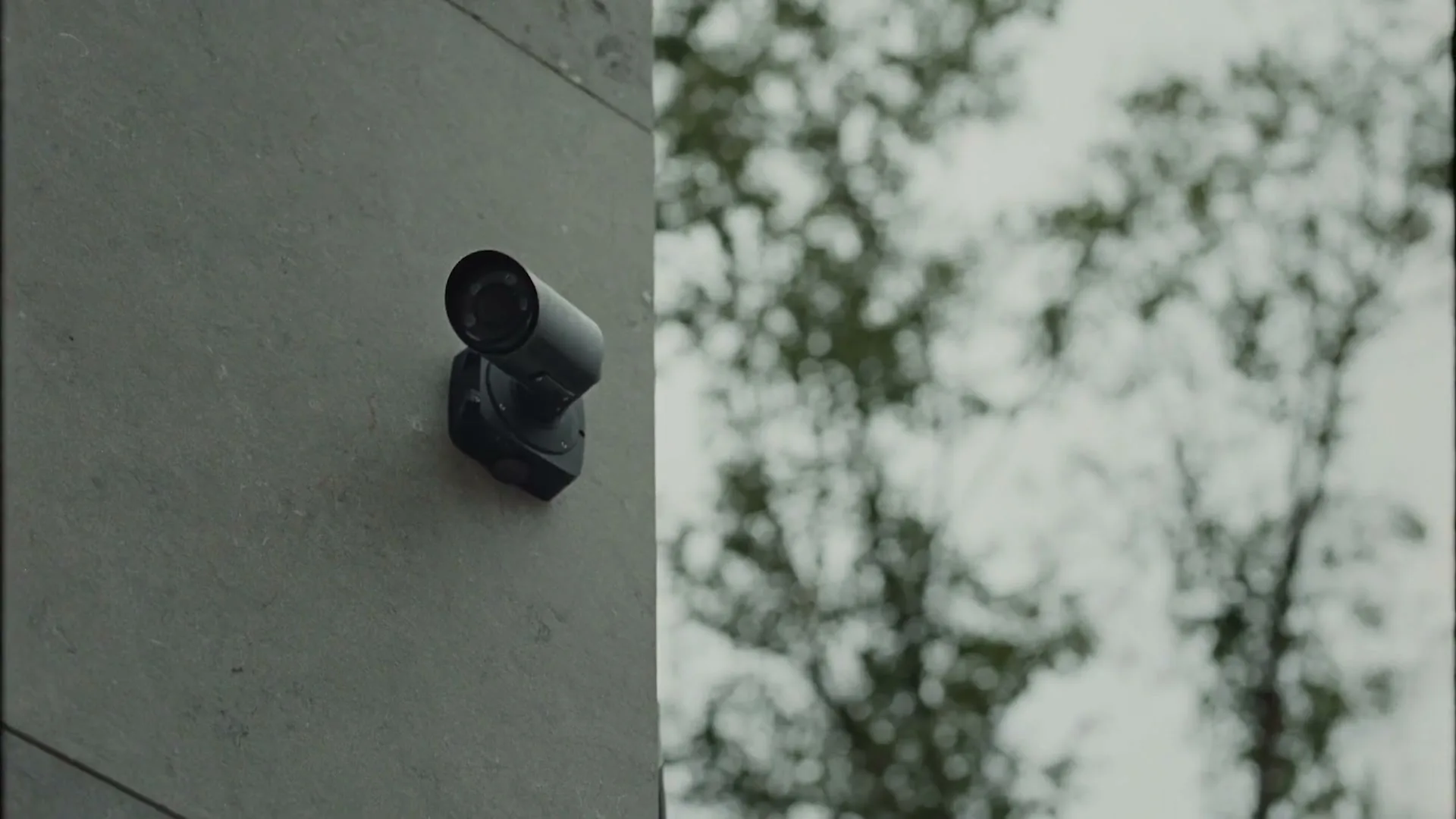A black security camera is mounted on a grey wall, with blurred green trees and a bright sky in the background.