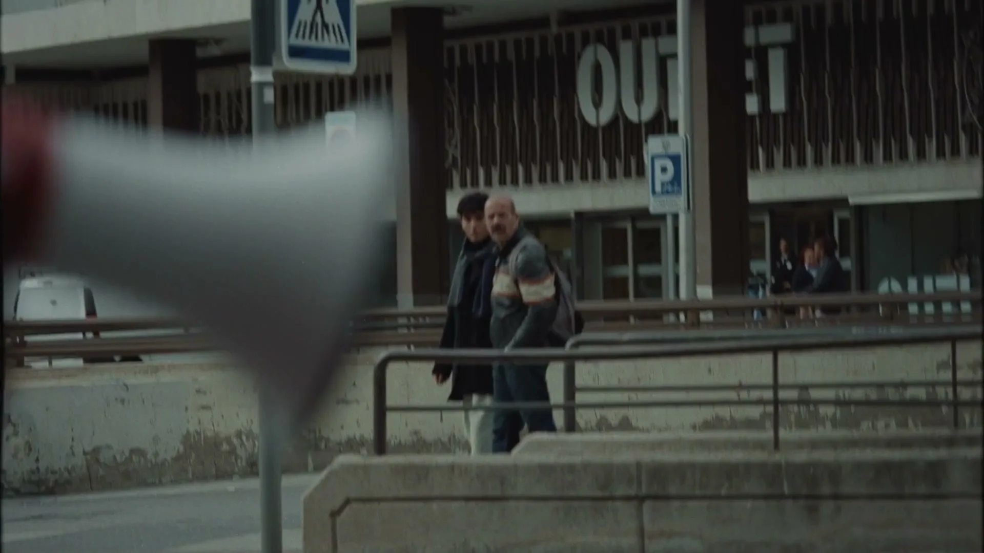 Two men walk past a building with "OUTLET" written on it, as a megaphone is blurred in the foreground, on a city street.