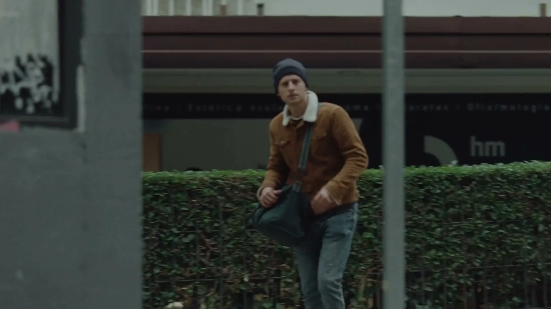 A man in a brown jacket and blue beanie walks on a city street, looking around nervously, with "No to war!" text on screen.