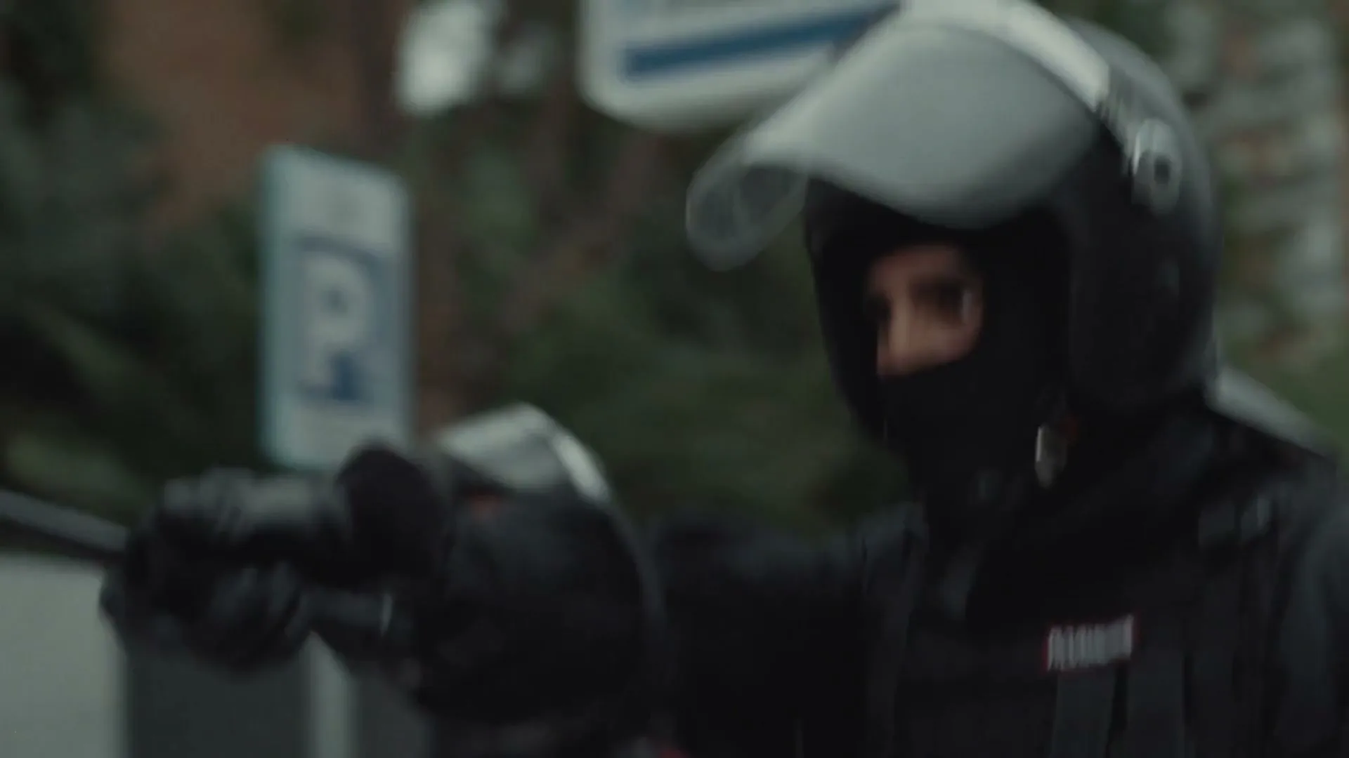 A group of riot police in black uniforms and helmets confront and arrest protesters on a city street during the day.