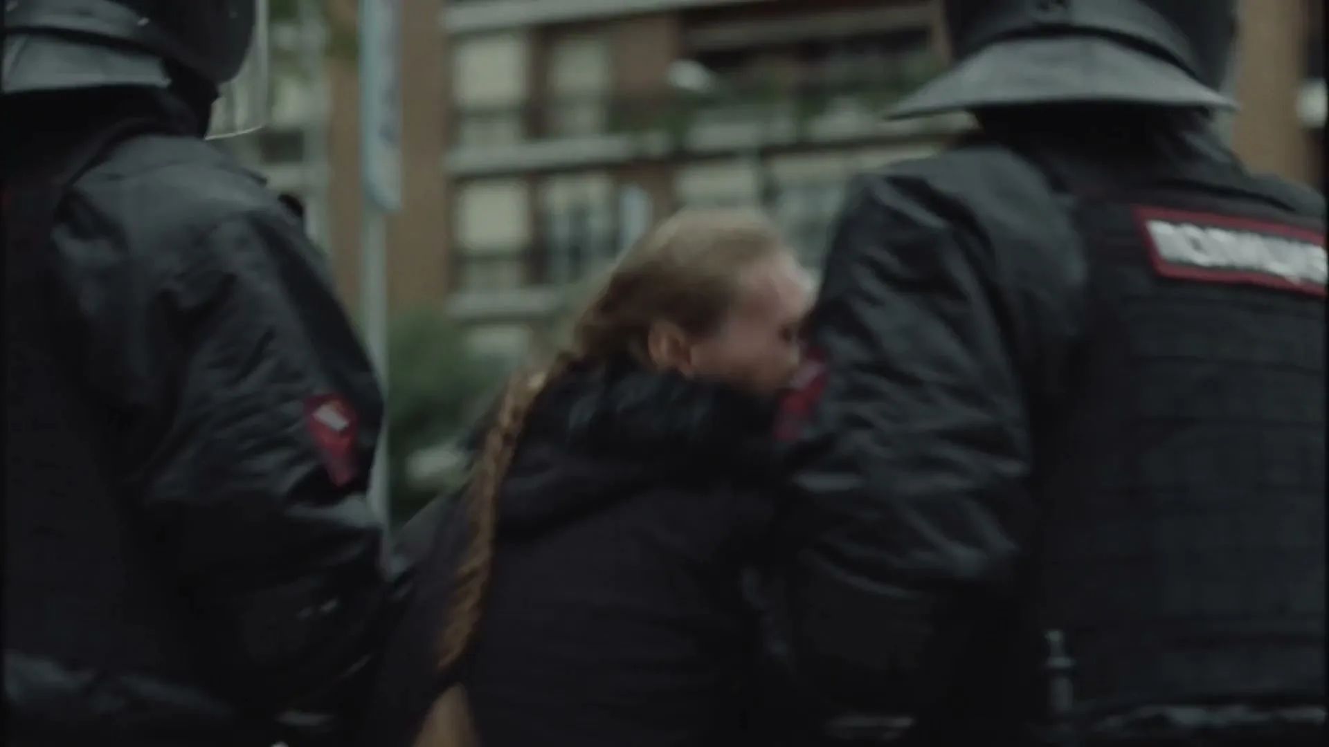 A man in a black jacket is pinned to the ground by an unseen force, while a woman with long hair is forcefully led away by two police officers in riot gear on a city street.