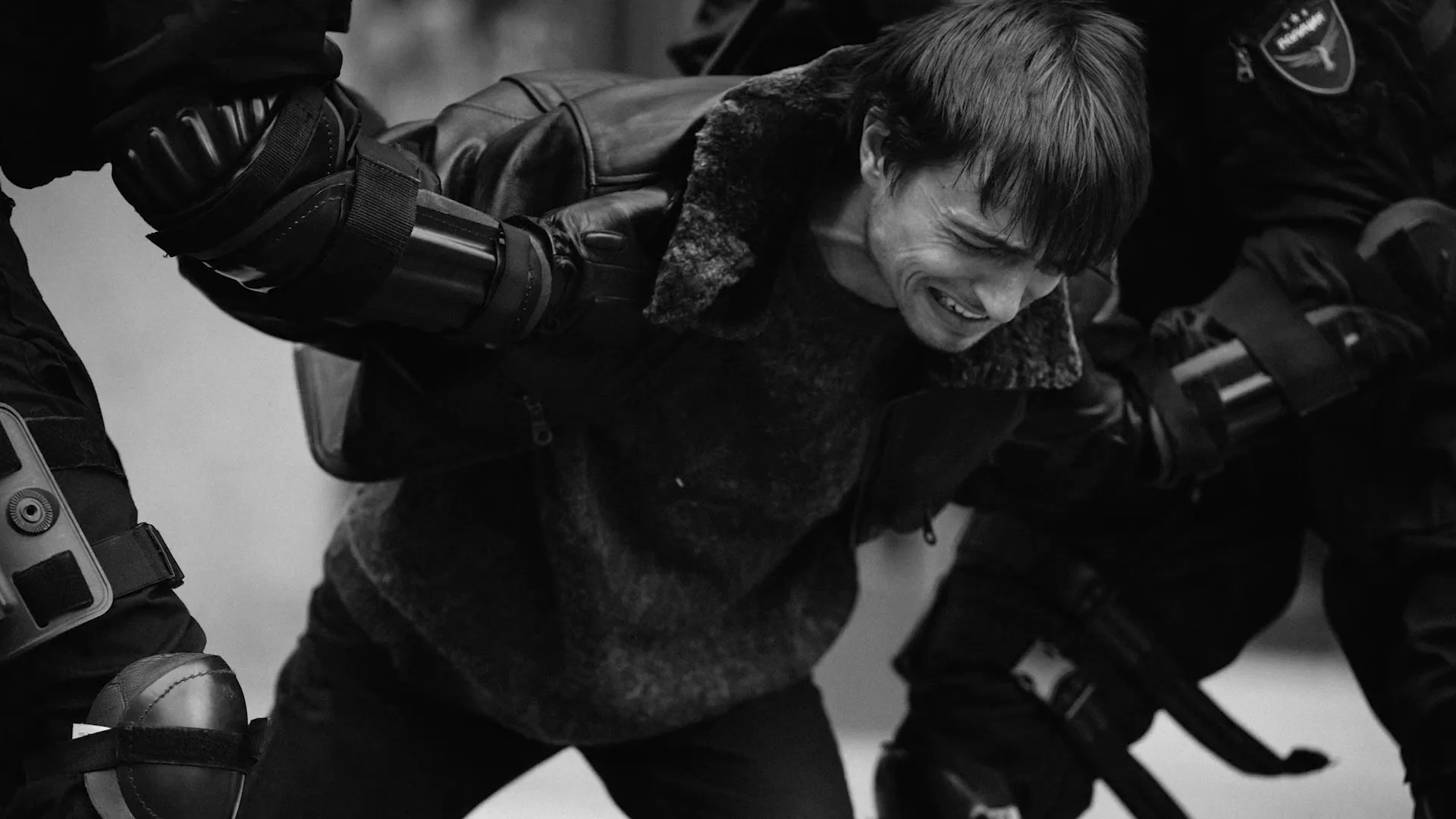 A man in a leather jacket is being forcefully restrained by two people in tactical gear, his face contorted in pain, in a black and white shot.