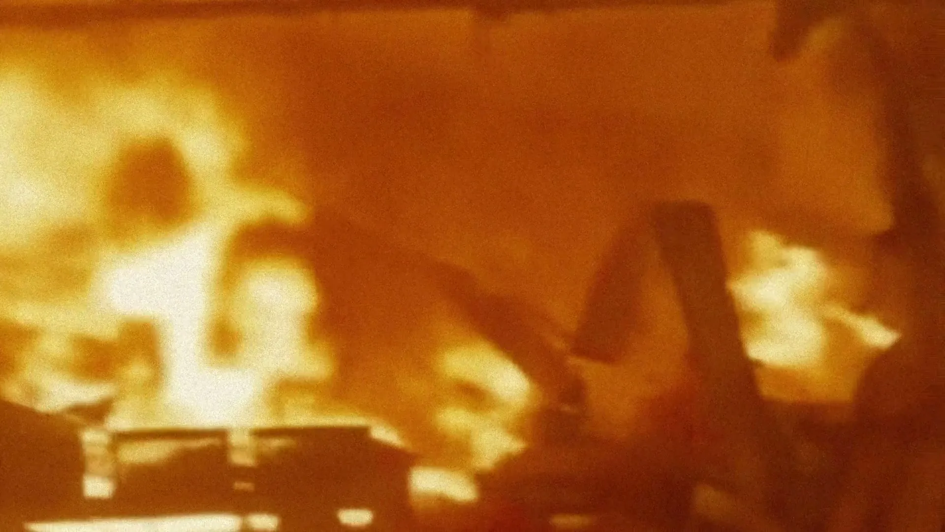 A blurry, grainy shot shows a building engulfed in bright orange flames and smoke, with indistinct shapes of debris or structures visible within the inferno.