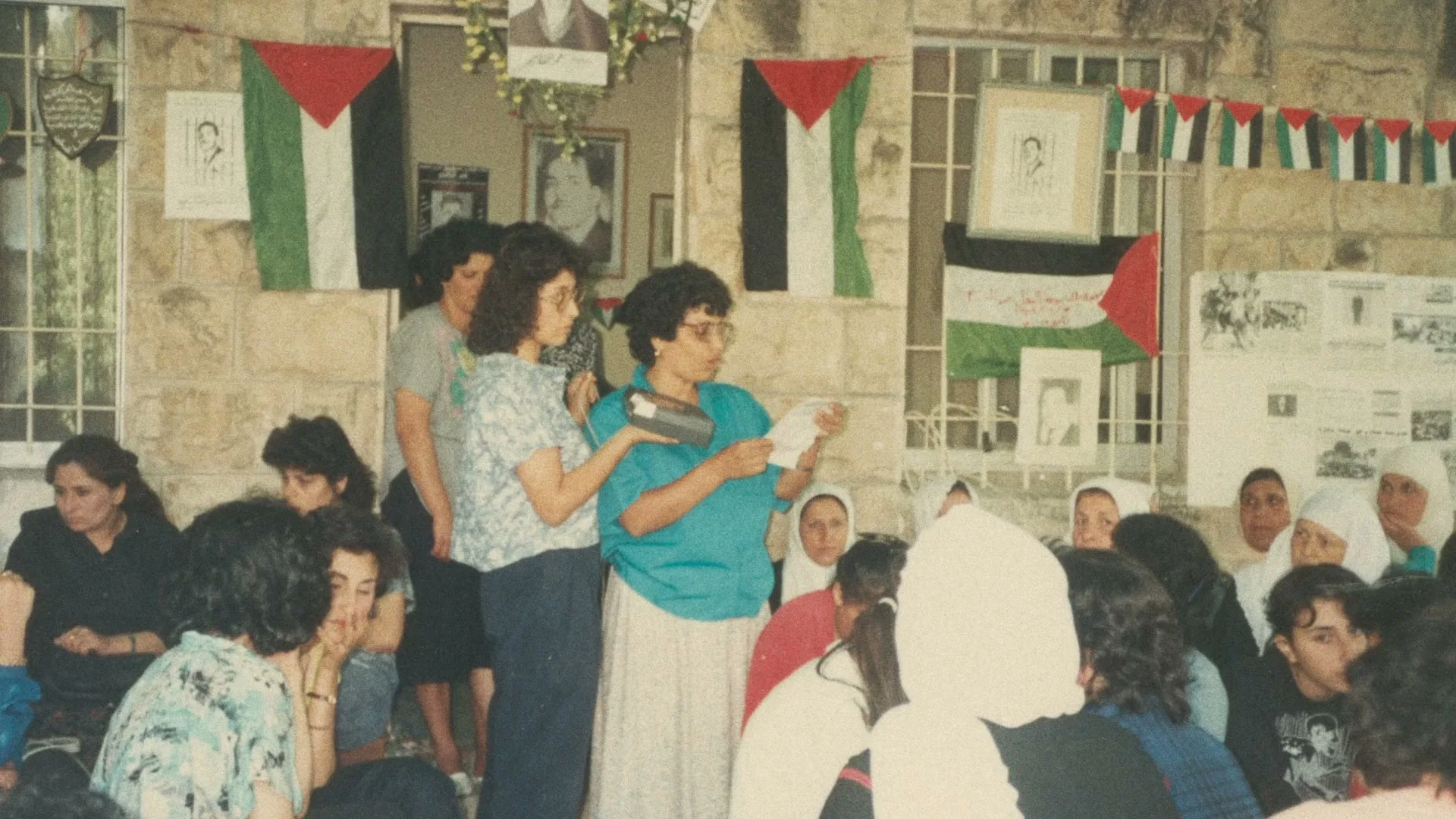 A group of Palestinian women gather, some holding flags and documents, in a stone building adorned with Palestinian flags and portraits, during what appears to be a community meeting or event.