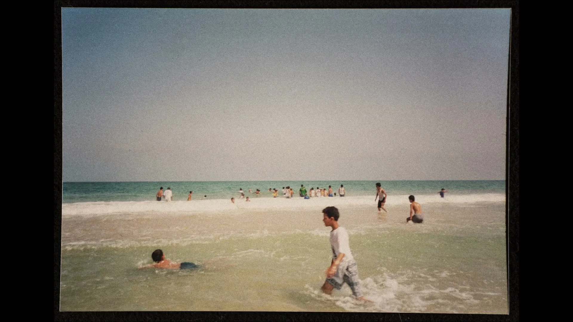 A group of children and teens play in the shallow waters of a beach on a bright, sunny day.