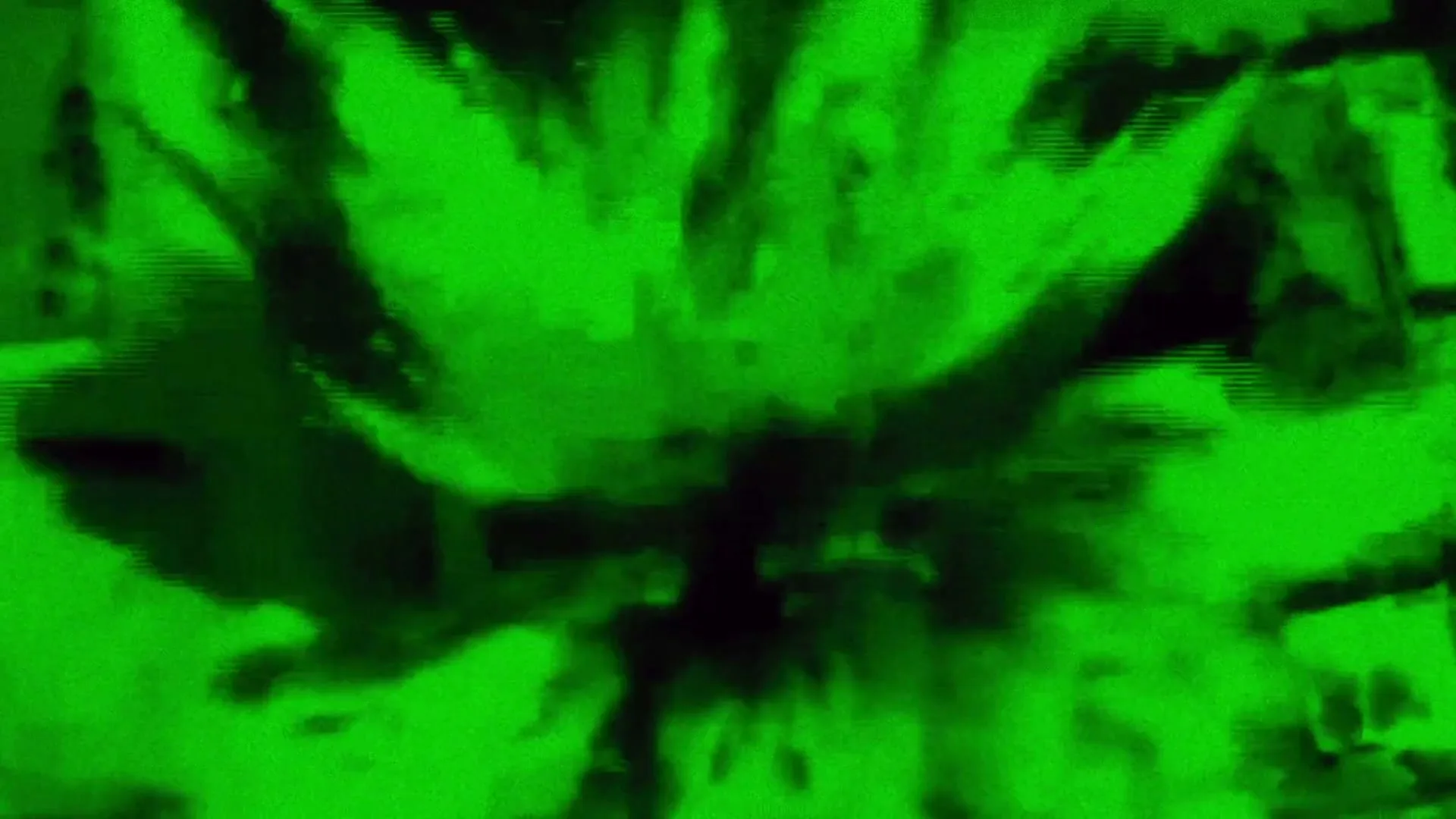 An aerial view of a green-tinted landscape with buildings and trees, seen through a night vision scope, as the view rapidly zooms into a dark, blurry center.