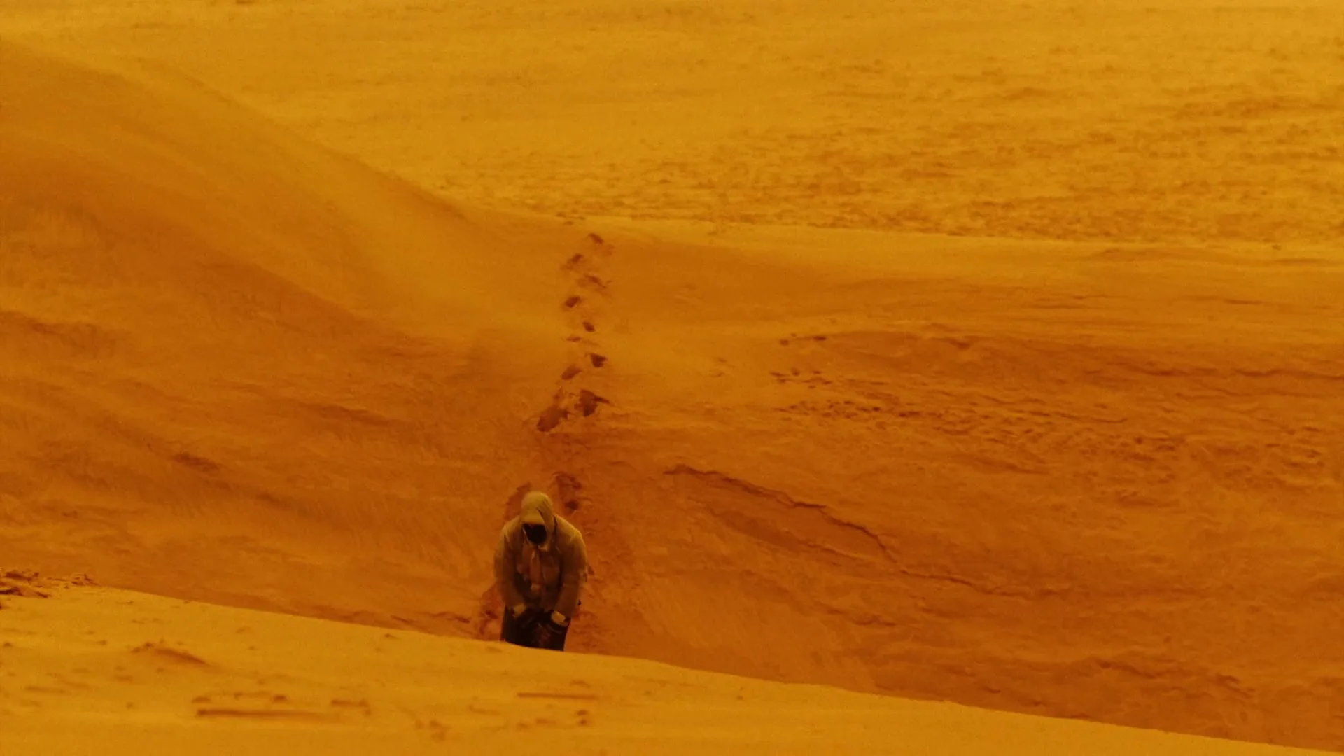 A person in a hooded jacket walks through a vast, desolate, orange-hued desert, leaving footprints behind, as the camera pulls back to reveal another person in the distance.