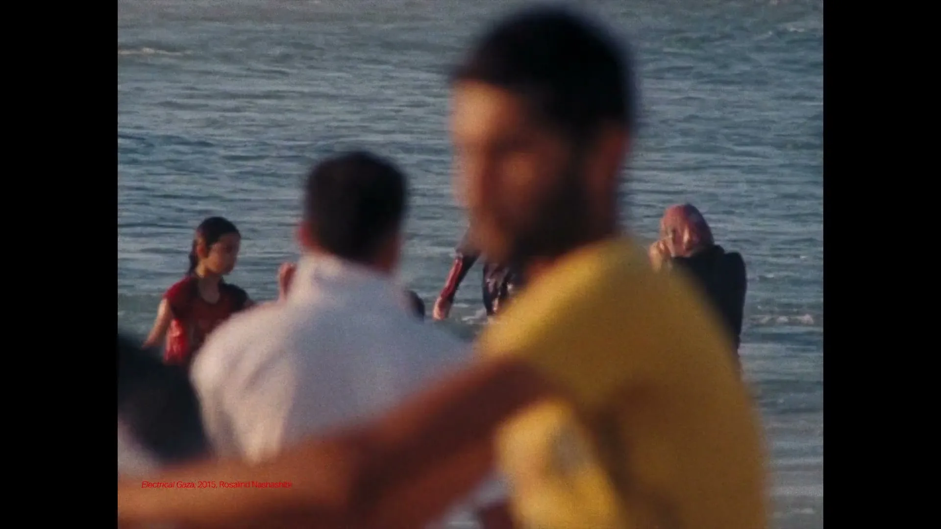 A group of people, including children, are seen wading and playing in the ocean water on a sunny day, with some figures blurred in the foreground.