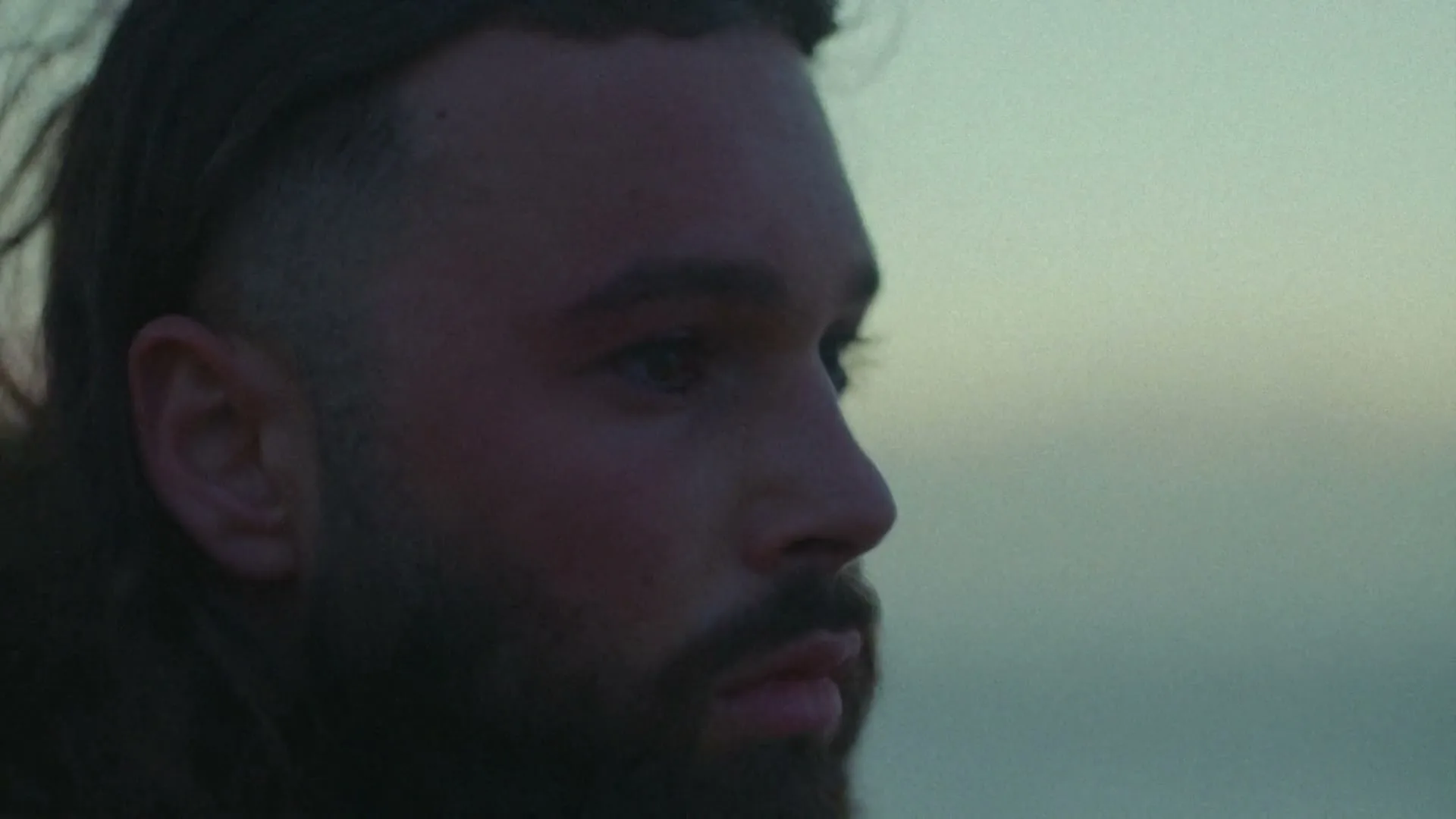 A young man with a beard and a fade haircut looks off into the distance with a serious expression, against a soft, out-of-focus background.
