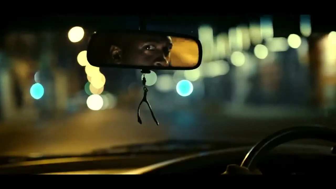 A man's eyes are reflected in a car's rearview mirror, looking intently at something off-screen, with blurred city lights in the background at night.