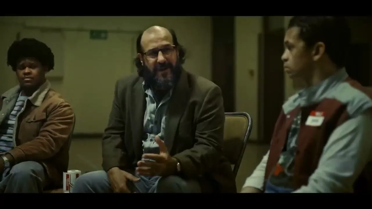 Three men sit in a circle in a dimly lit room, with the man in the center speaking and gesturing with his hands.