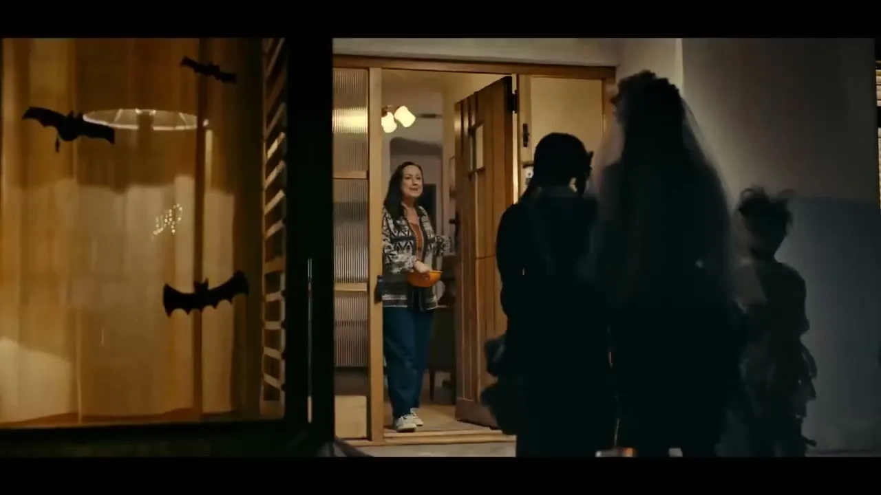 A woman in a patterned cardigan stands in a doorway, holding a bowl of candy, as three children in Halloween costumes approach her at night.