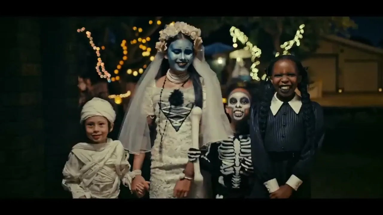 Four children in Halloween costumes, including a mummy, a zombie bride, a skeleton, and Wednesday Addams, stand together at night, smiling and looking at the camera.