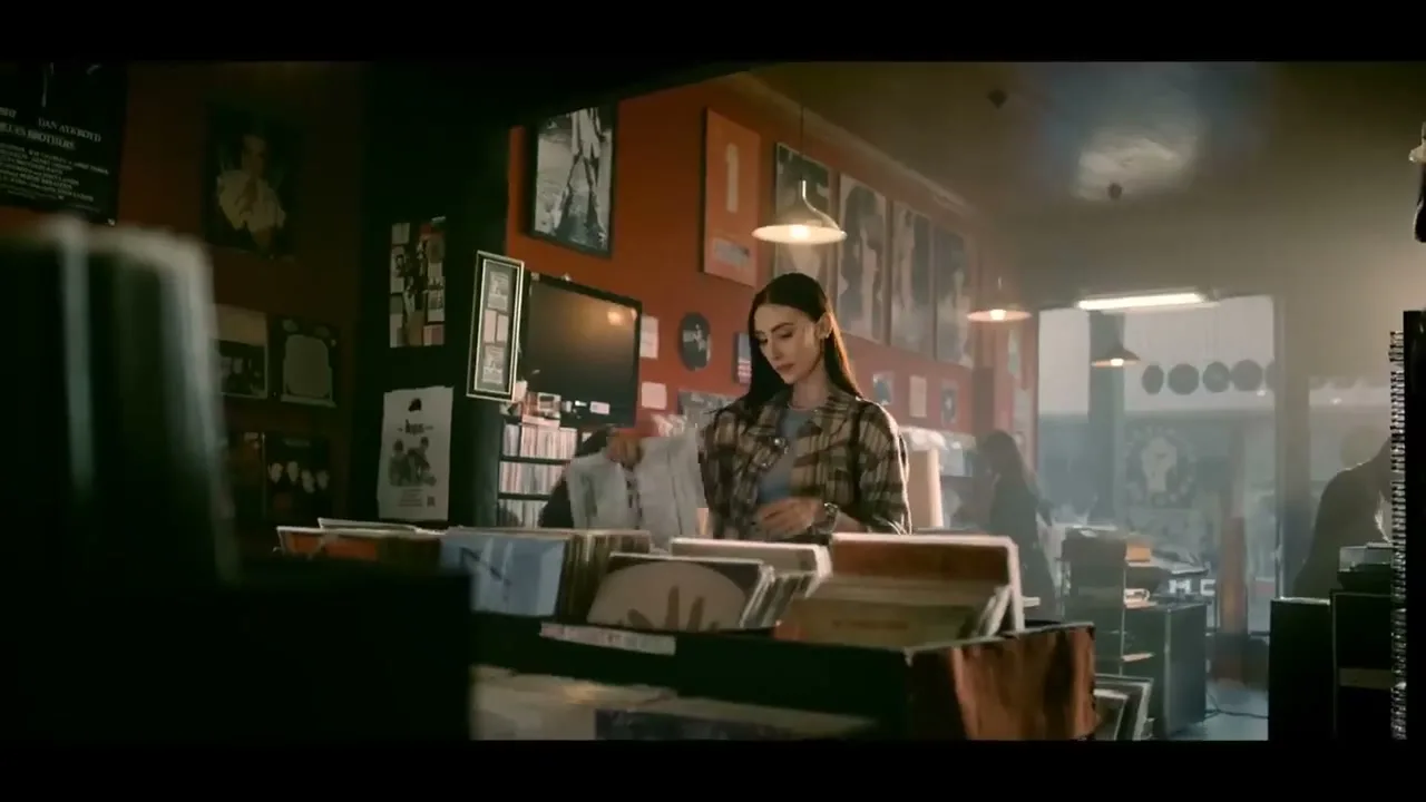 A young woman with long dark hair, wearing a plaid shirt, browses through vinyl records in a dimly lit record store.