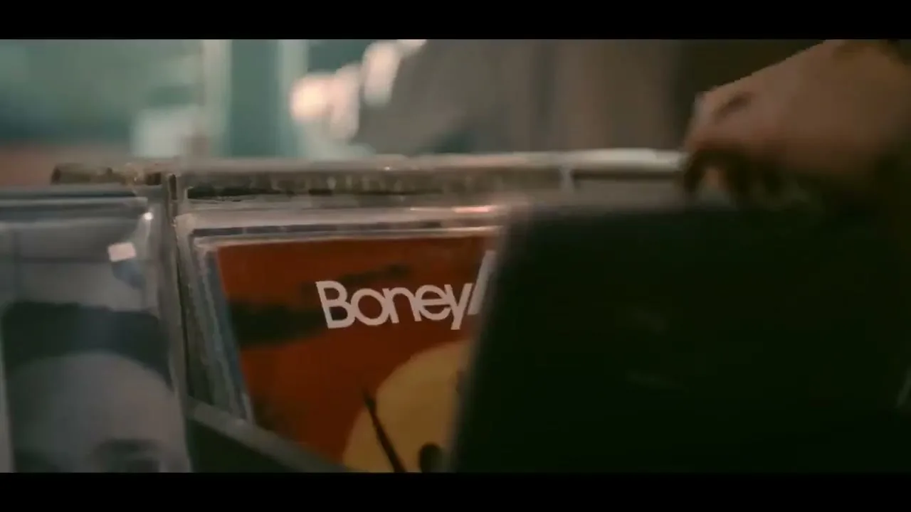 A person's hand flips through a collection of vinyl records in a dimly lit record store, revealing a "Boney M." album cover.