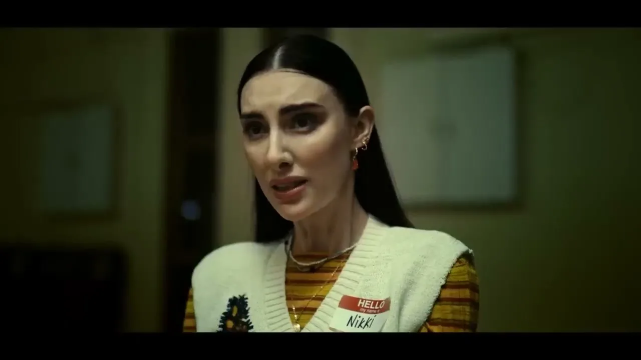 A pale woman with dark hair and a name tag looks concerned, wearing a striped shirt and a cream vest, in a dimly lit room.