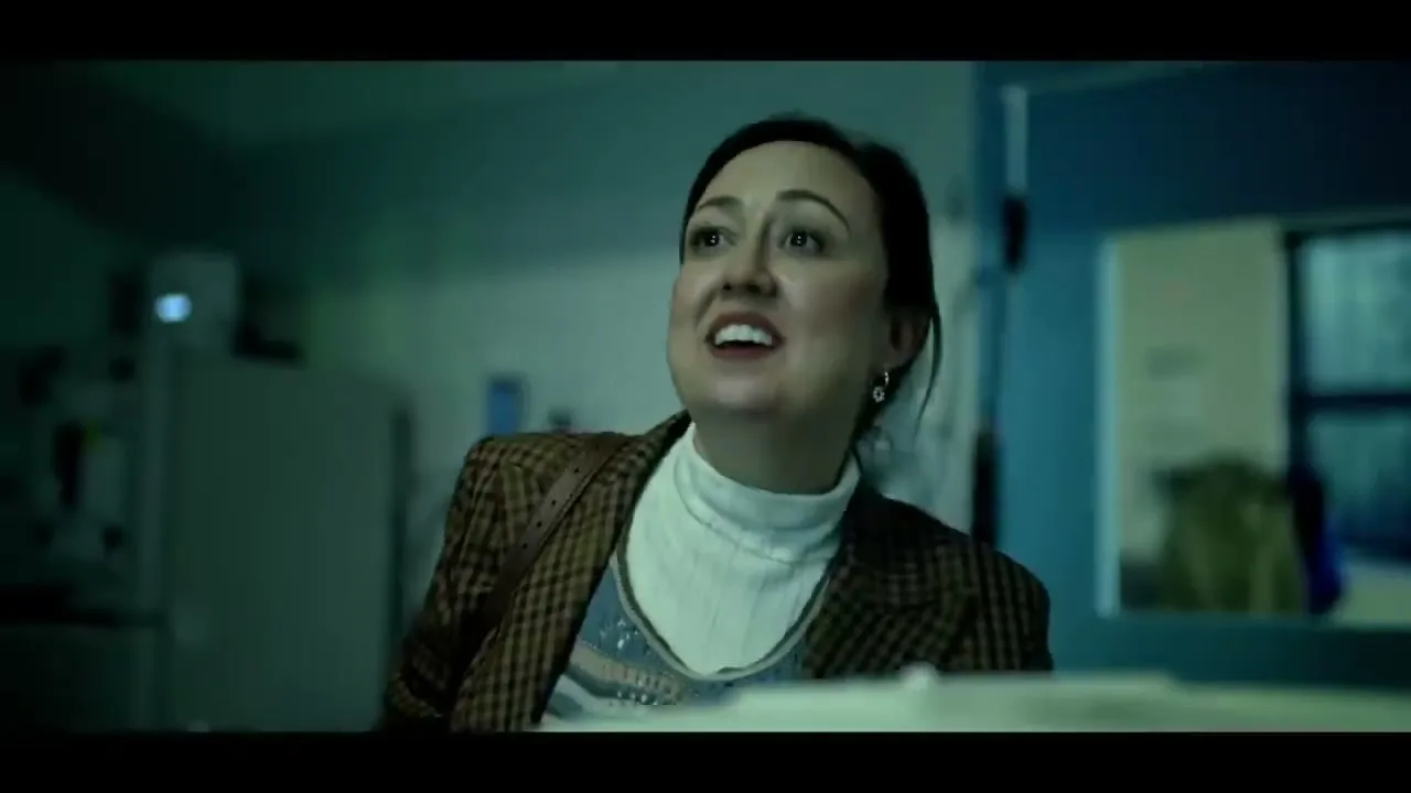 A woman in her 30s, wearing a brown plaid blazer and white turtleneck, looks up with a pained expression in a dimly lit office kitchen.