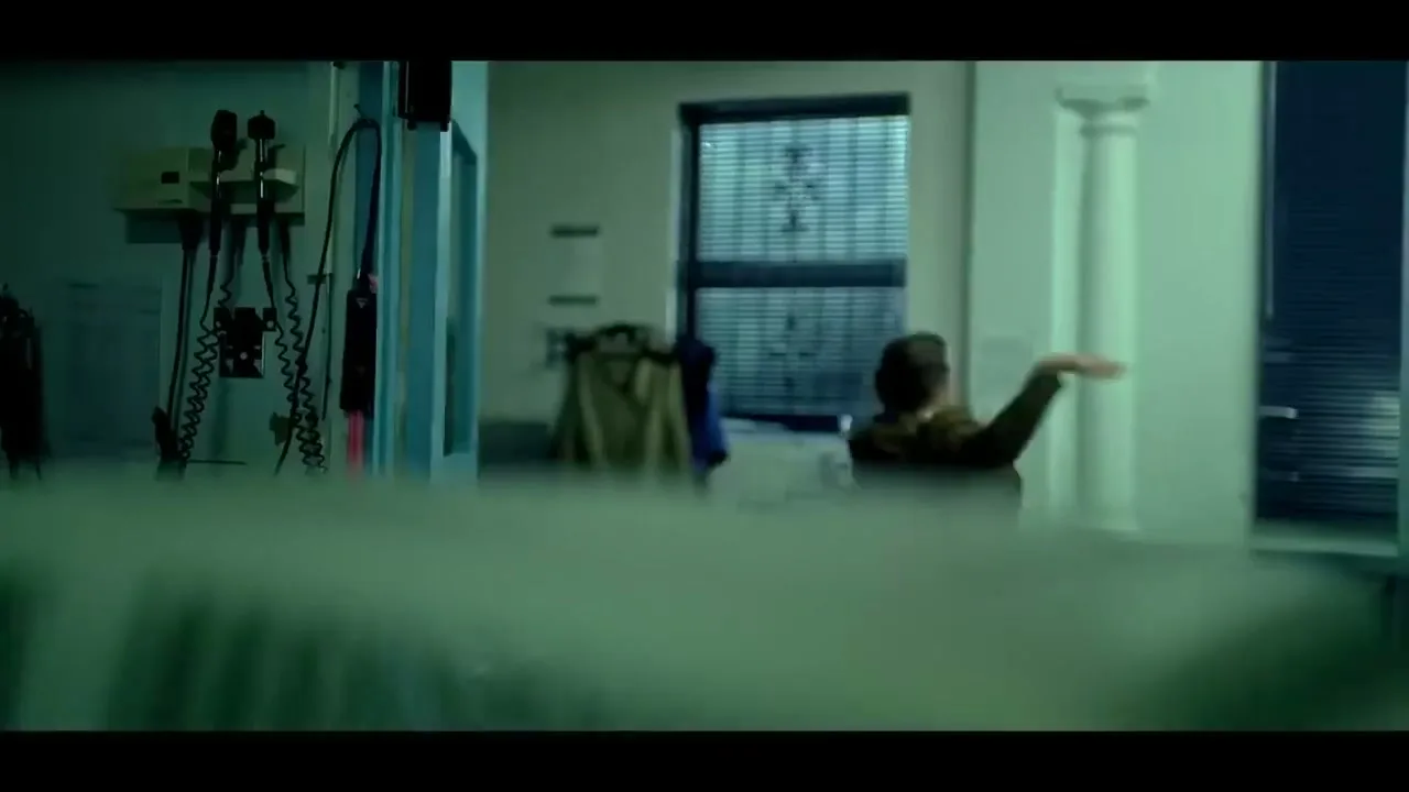A man in a brown jacket stumbles backward in a hospital room, seen from behind a blurred foreground.