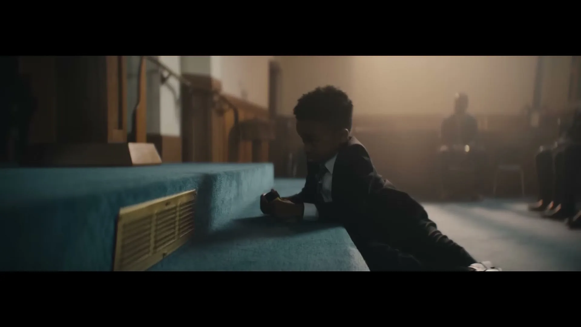 A young boy in a suit lies on the carpeted steps of a church altar, looking down at something in his hands.