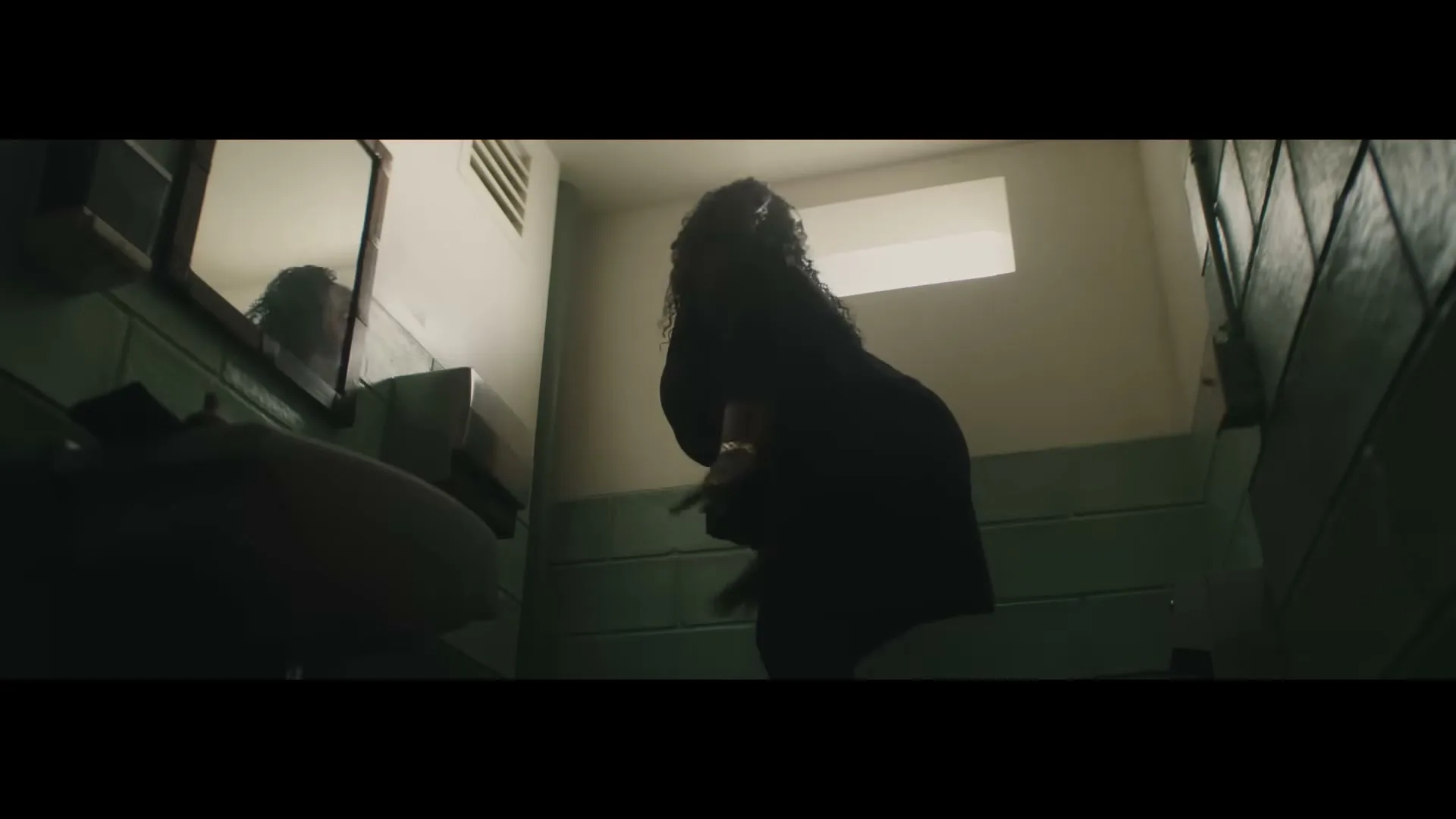 A woman in a dark dress is seen from behind, bending over in a dimly lit public restroom with green tiled walls.