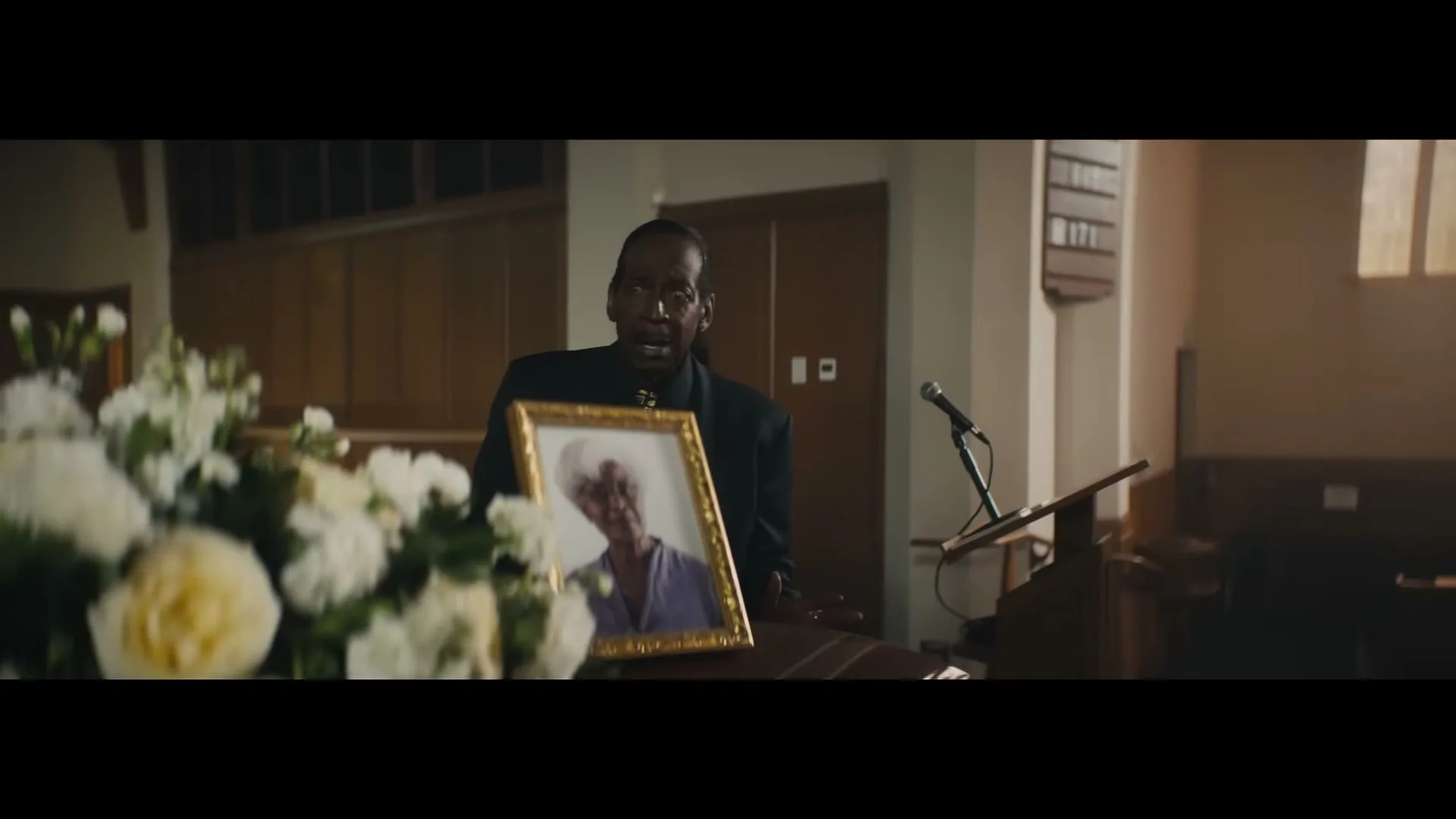 A man in a black suit stands at a podium with a framed photo of an older woman, looking emotional in a church.