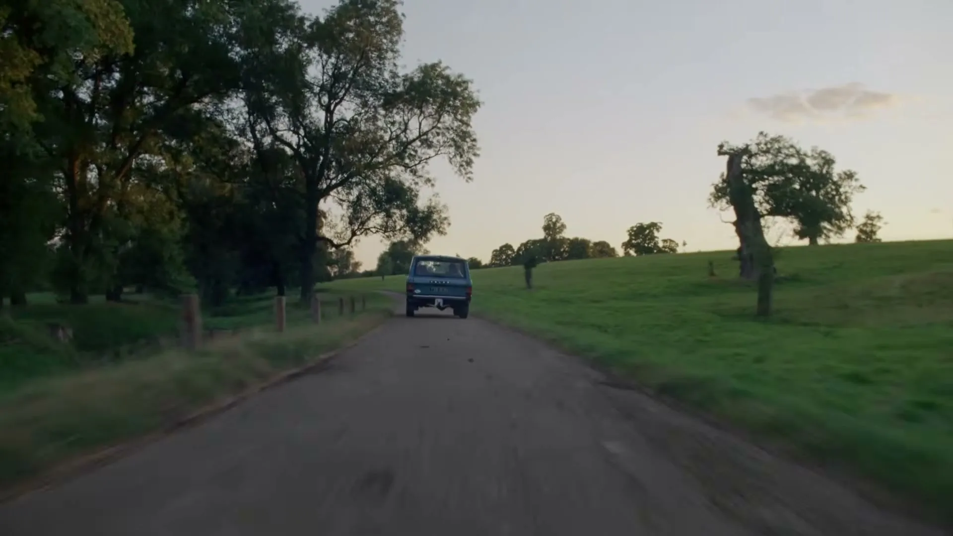 A blue vintage van drives away down a dirt road, surrounded by green fields and trees under a soft evening sky.