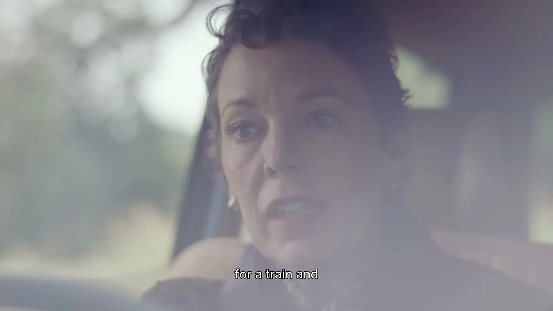 Olivia Colman, a woman in her 40s, is seen from the chest up, talking while driving a car on a sunny day.